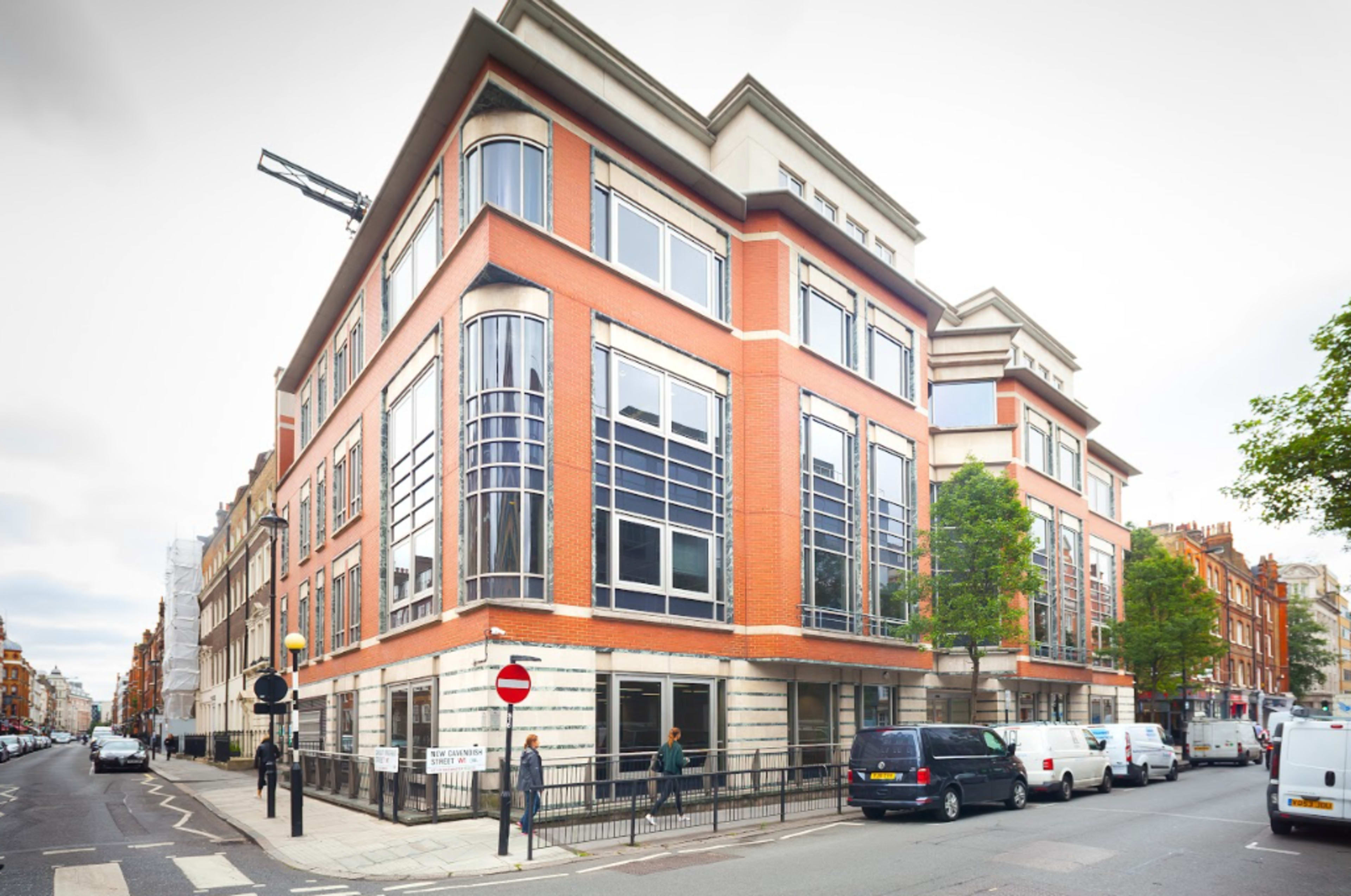 The image shows a modern brick building with large windows located at an intersection in an urban area, surrounded by parked vehicles and a pedestrian.