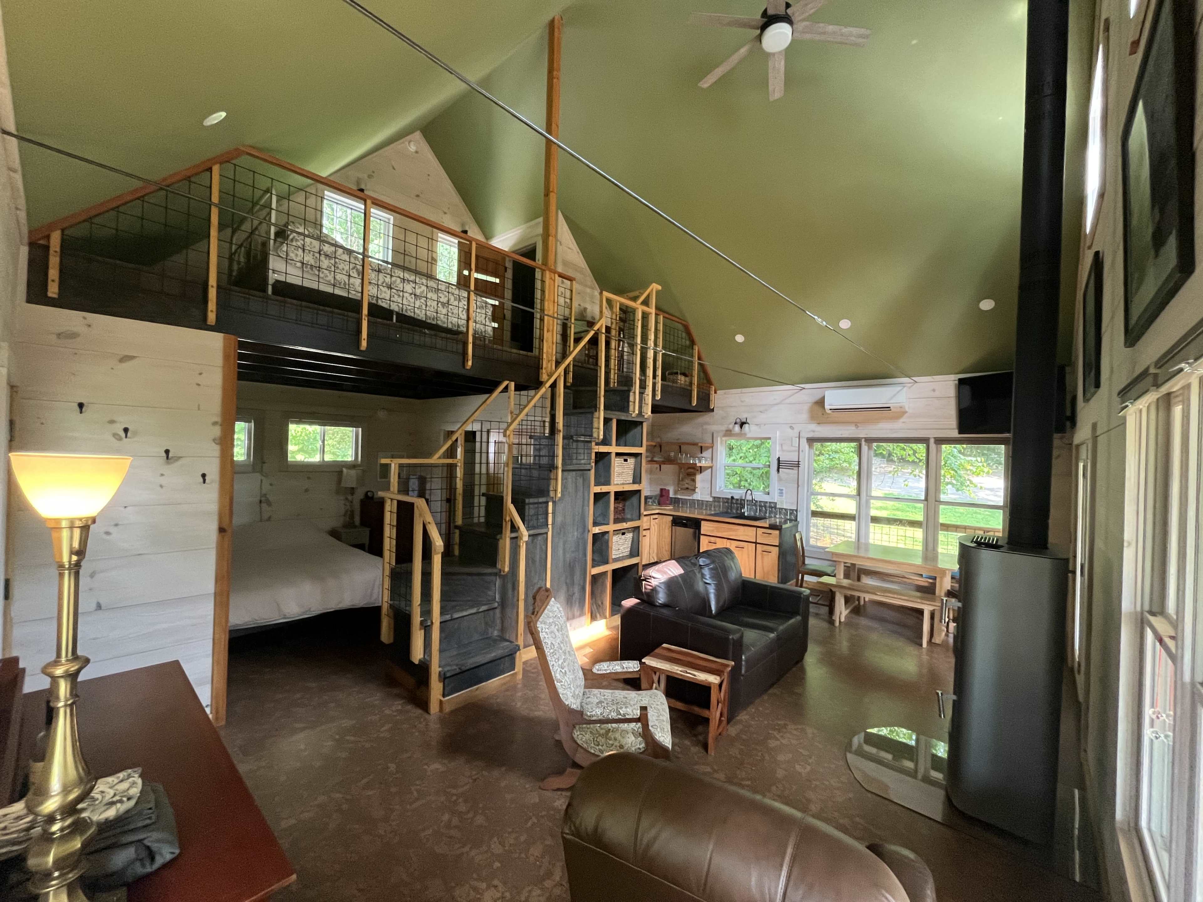 The image shows a spacious cabin interior featuring a seating area, a staircase leading to a loft, large windows, and modern amenities.