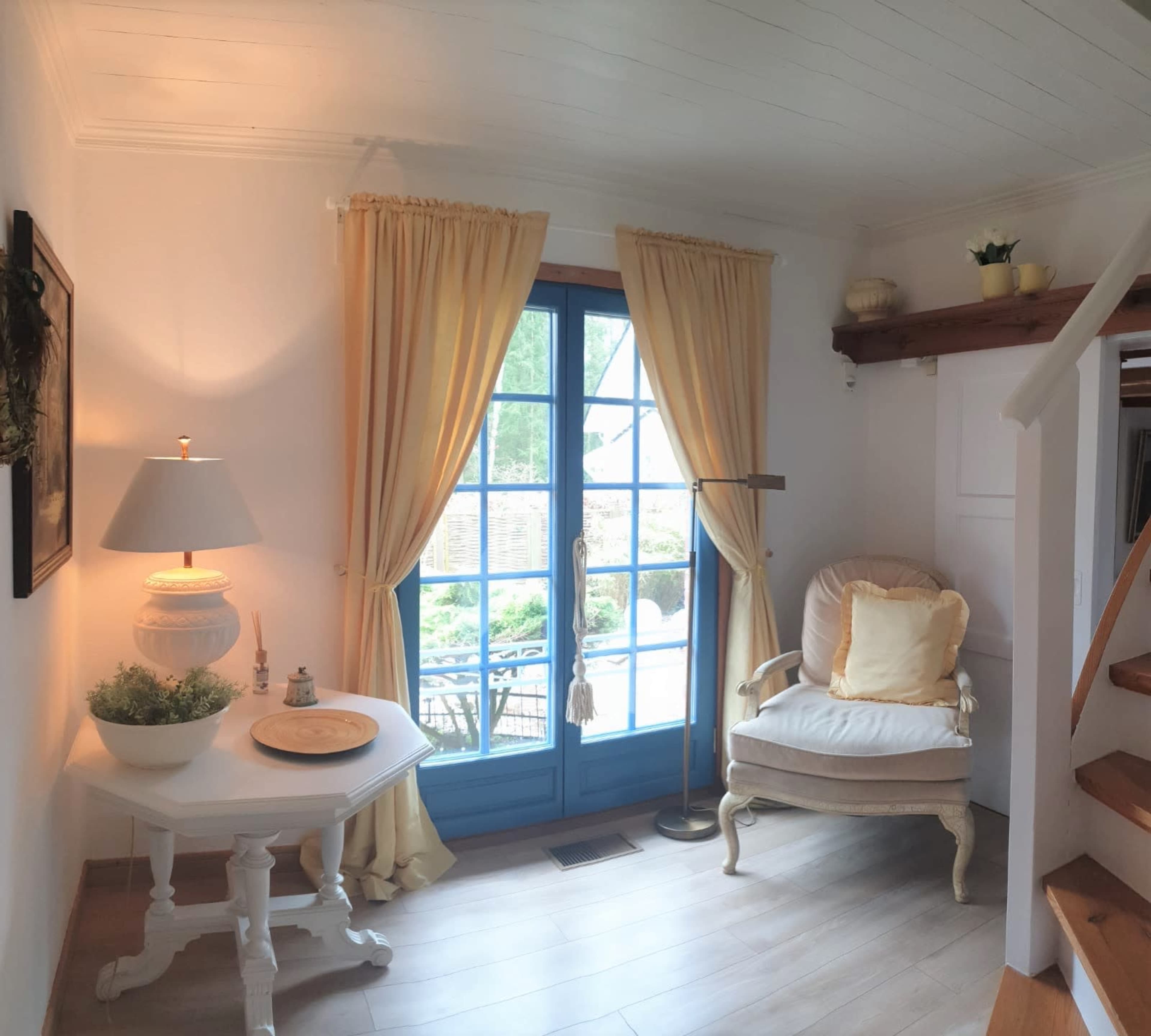 A cozy room features a white table, a cream-colored armchair, blue-framed windows with yellow curtains, and wooden stairs.