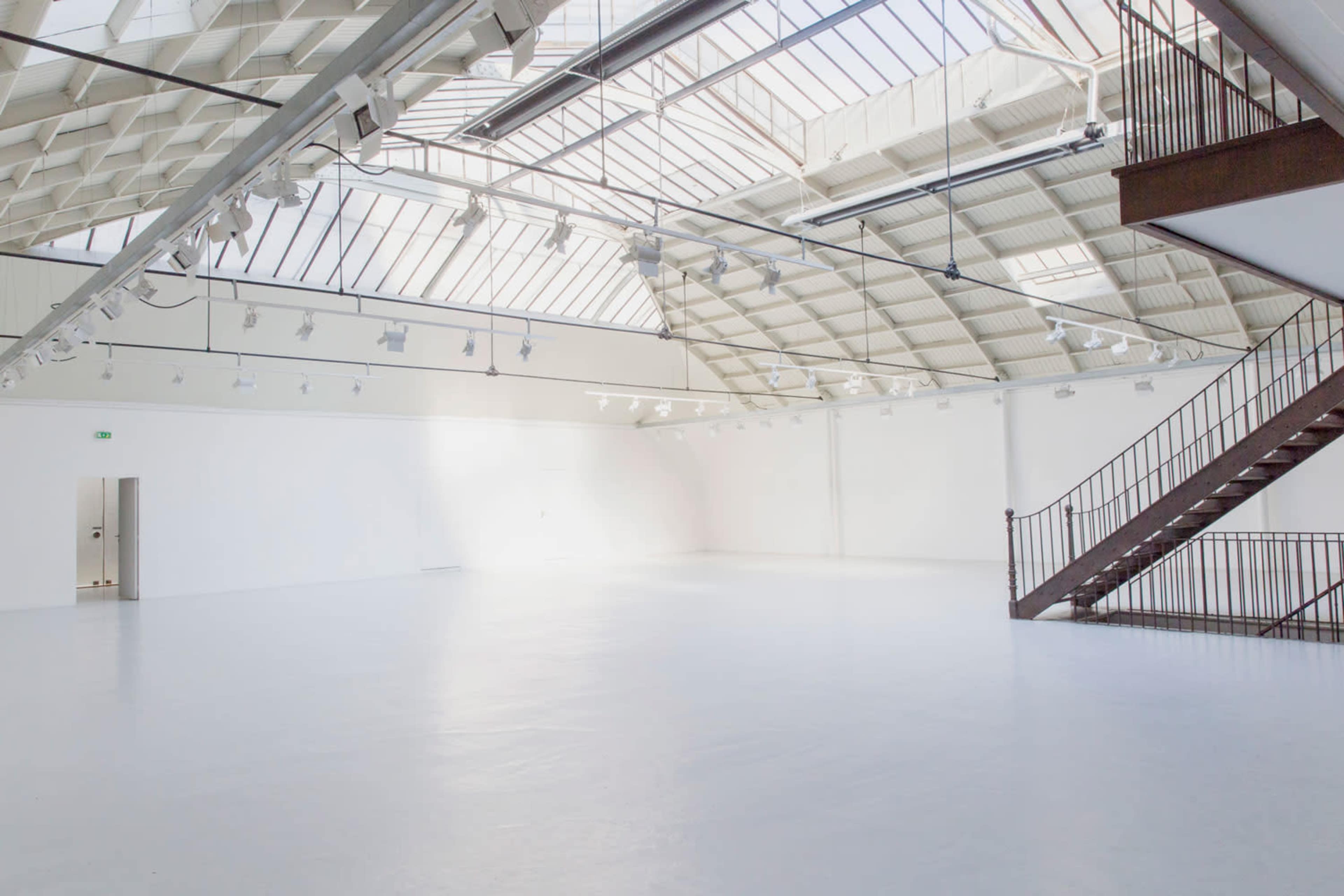 A spacious, well-lit art gallery with a high ceiling and a staircase in one corner.