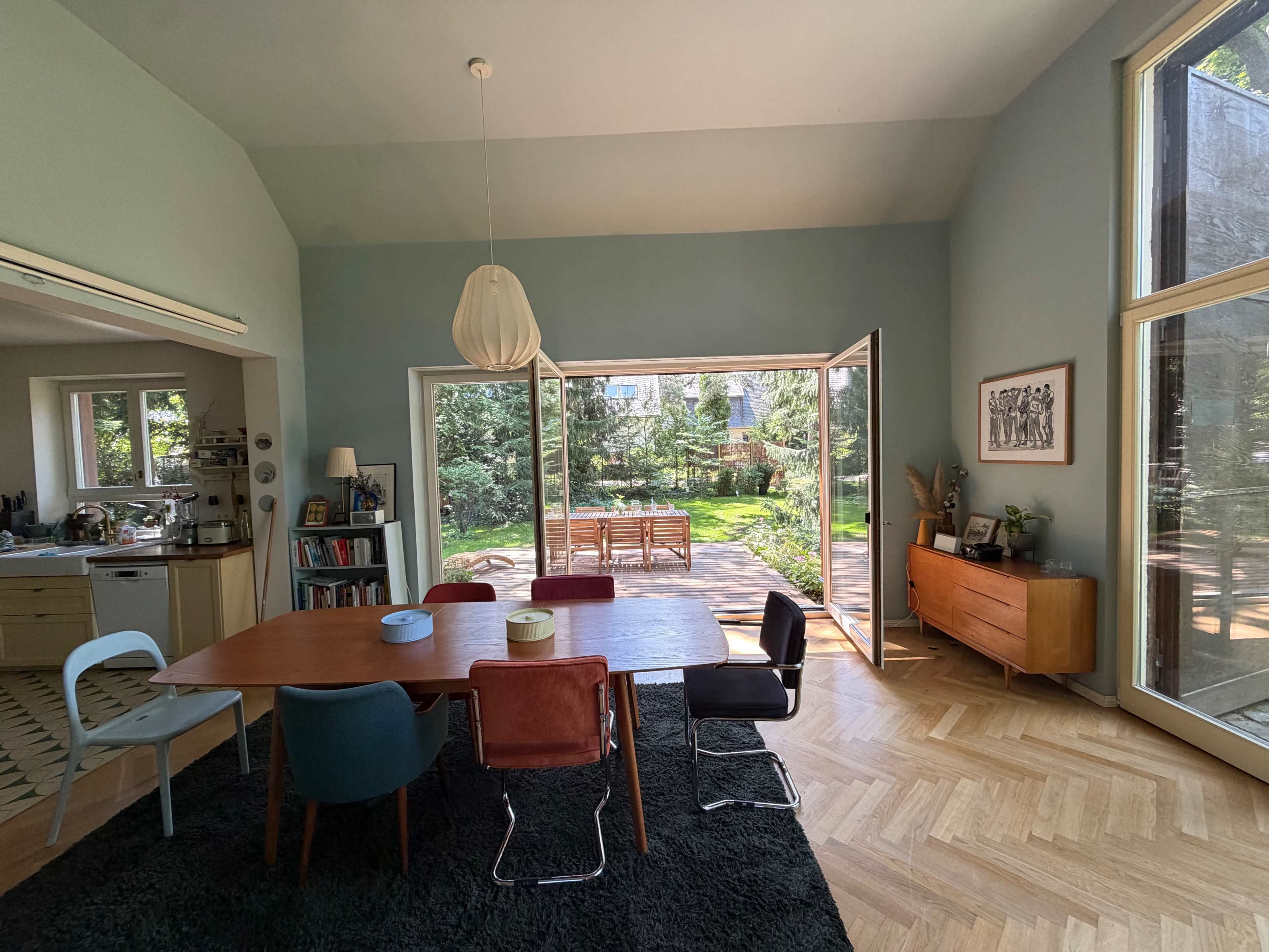 A modern dining area features a large table surrounded by chairs, with large glass doors opening to a garden outside.
