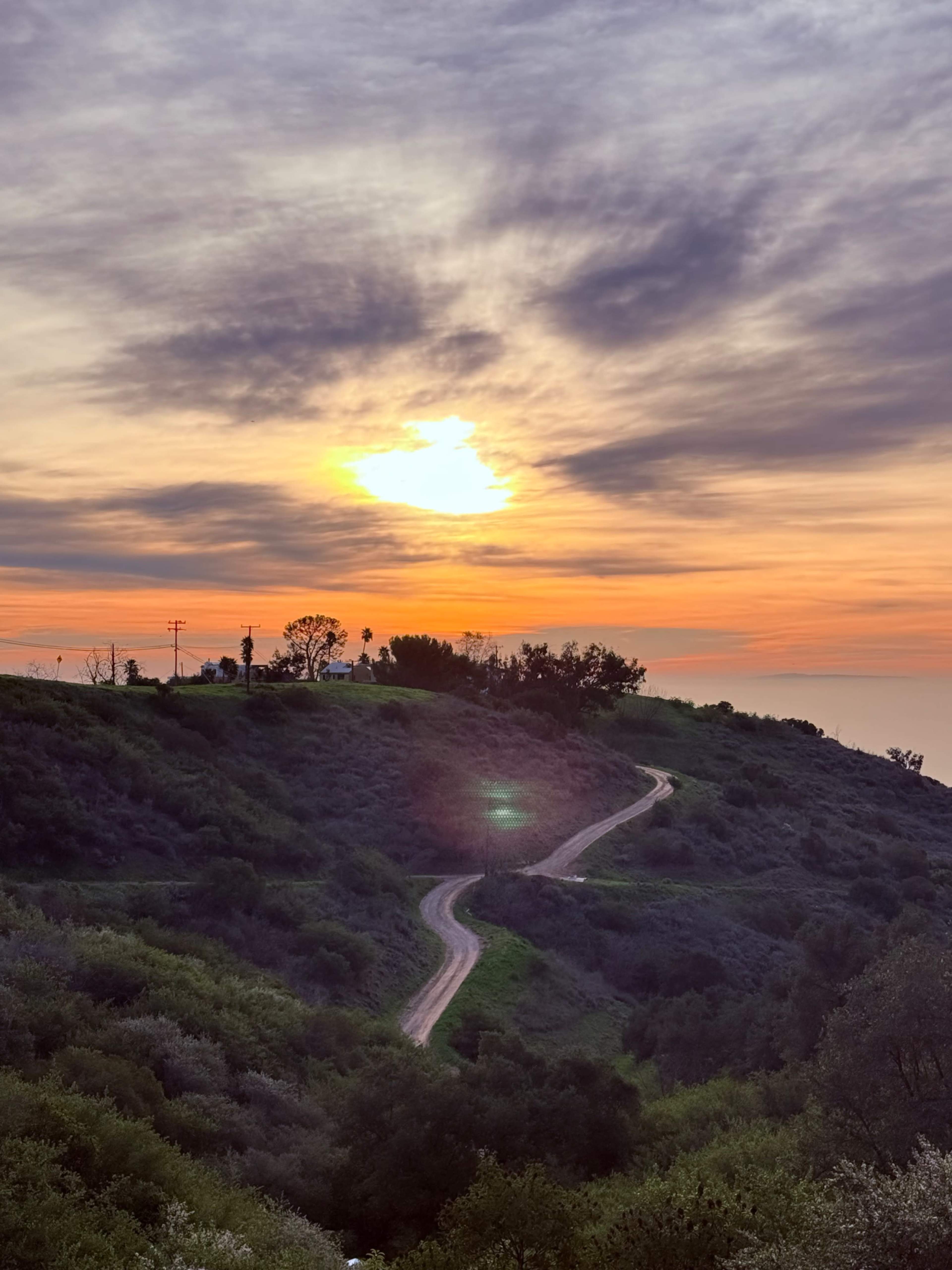 A winding dirt road leads along a hillside under a colorful sunset sky.