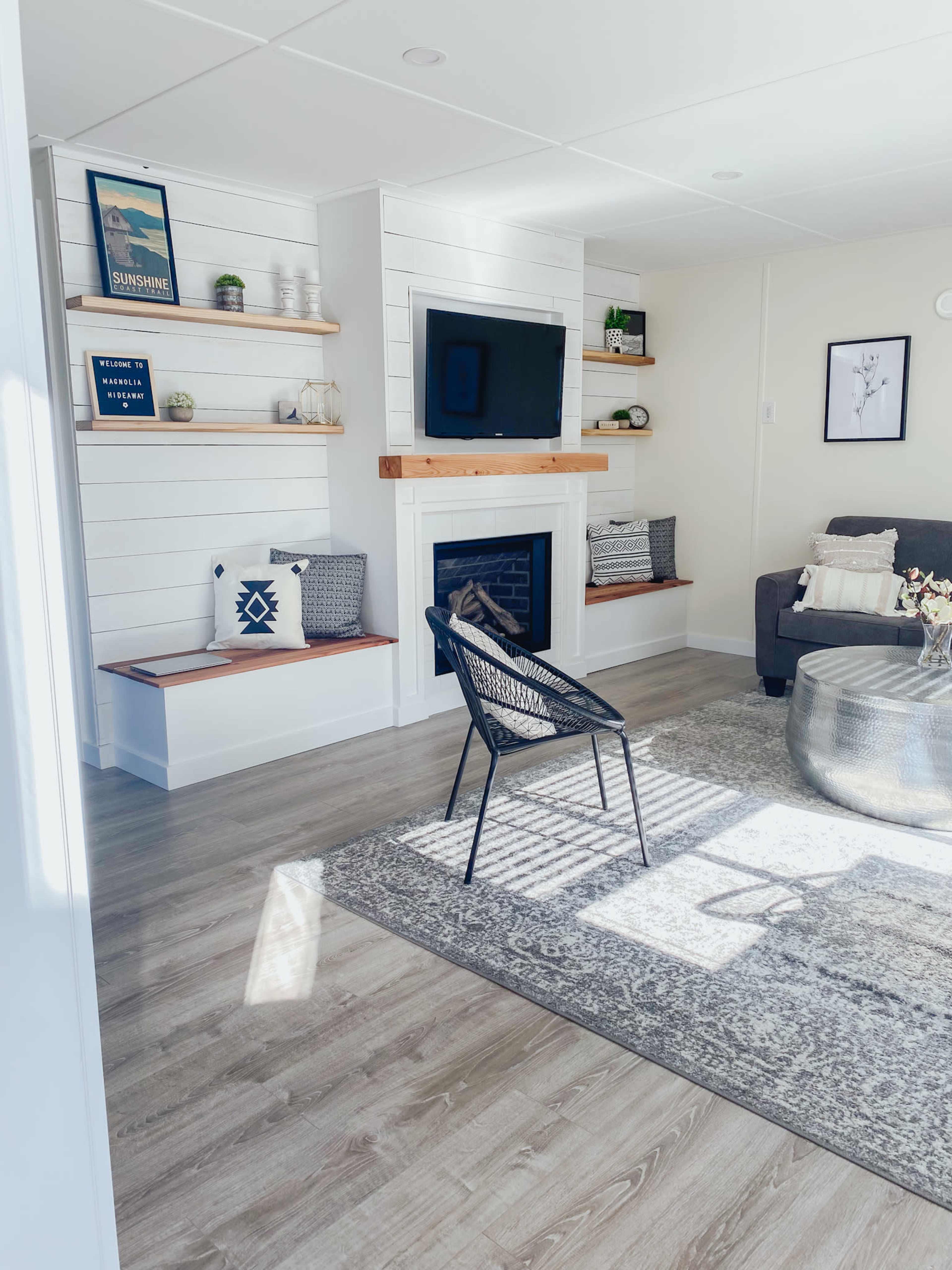 The image shows a modern living room with a television mounted above a white shiplap fireplace, flanked by shelving, a gray sofa, and a round coffee table on a patterned rug.