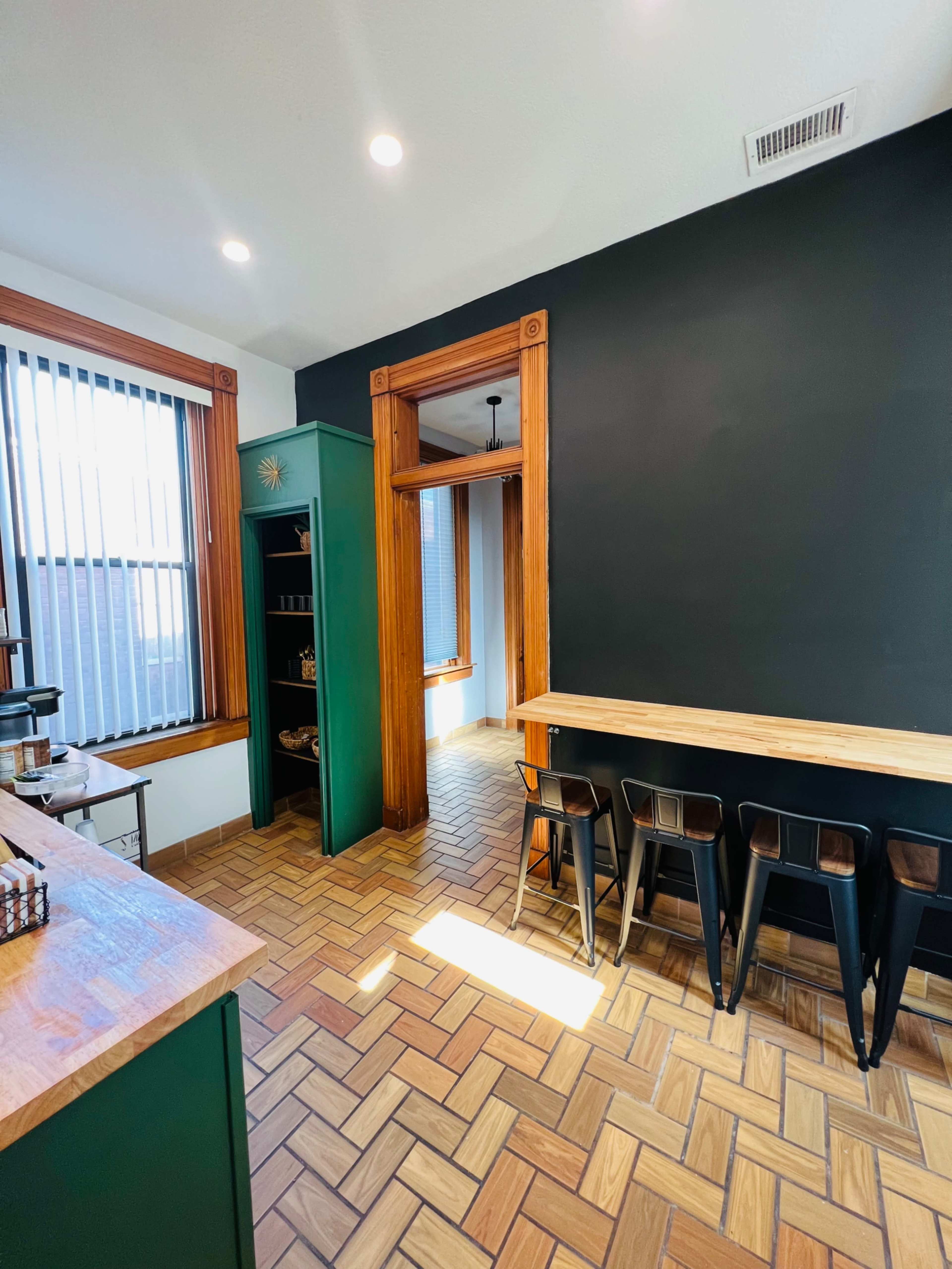 The interior of a café featuring wooden flooring, green cabinetry, and a countertop with black stools against a dark wall.