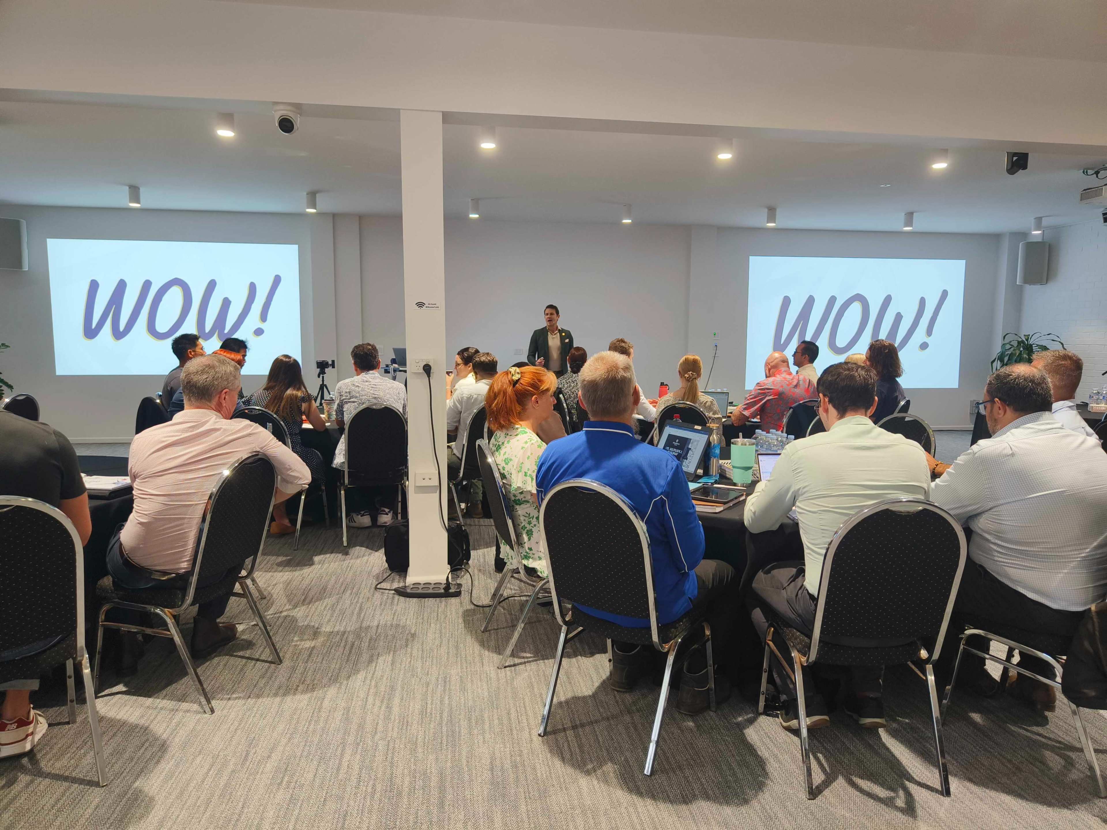 A meeting is taking place in a room with attendees seated at tables, facing a presenter in front of screens displaying the word "WOW!".