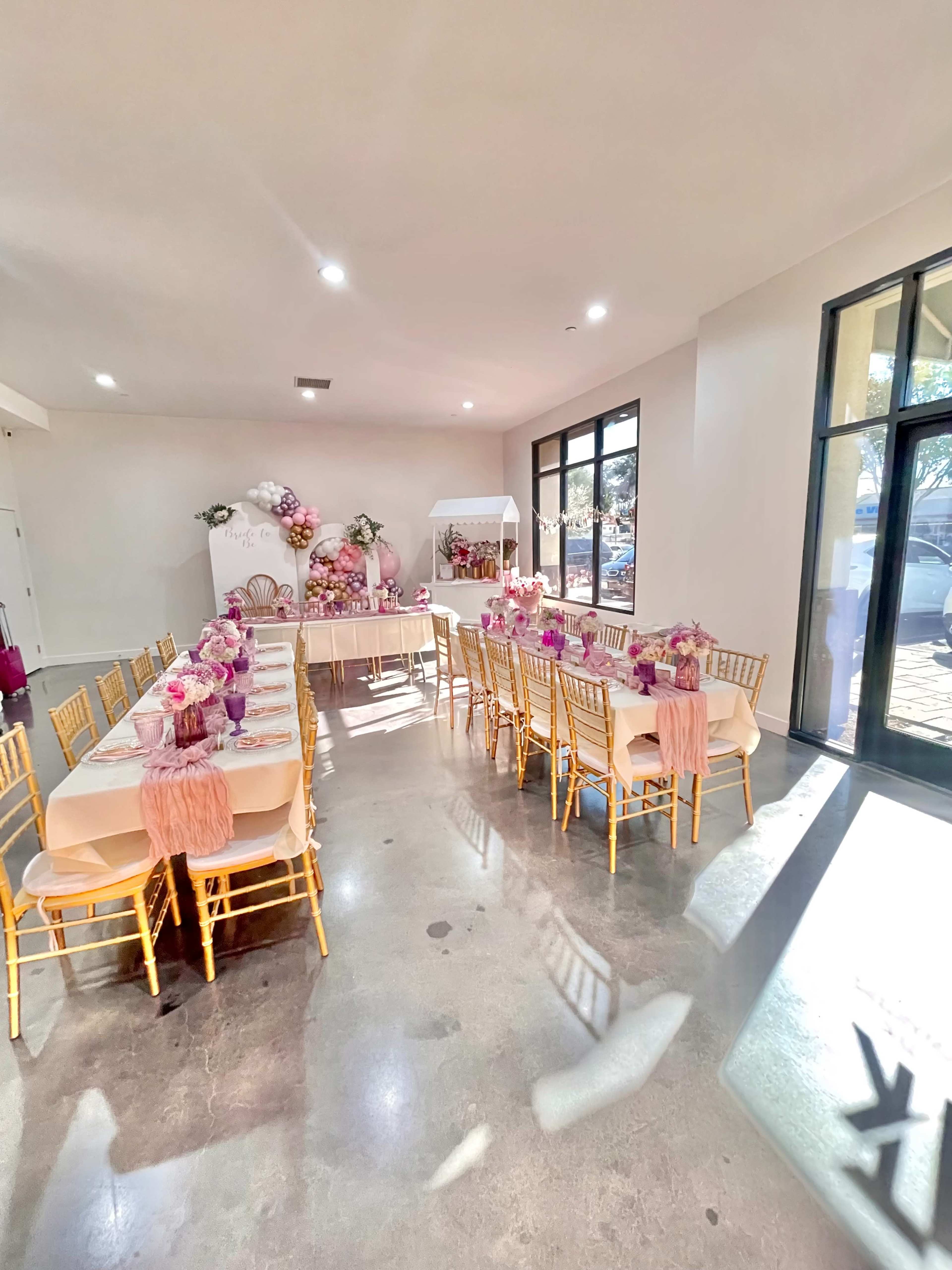 The image shows a decorated event space with tables set for a gathering, featuring pink floral arrangements and gold chairs.