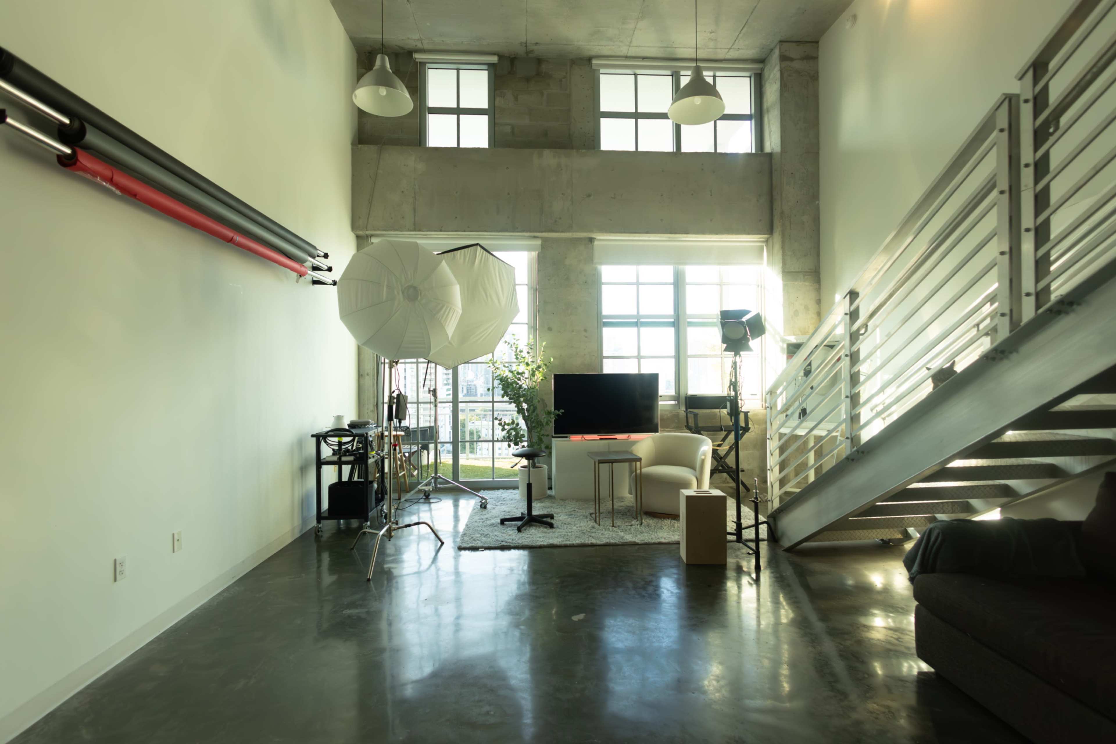 The image depicts a spacious, industrial-style room featuring large windows, photography equipment, and furniture arranged in a modern layout.