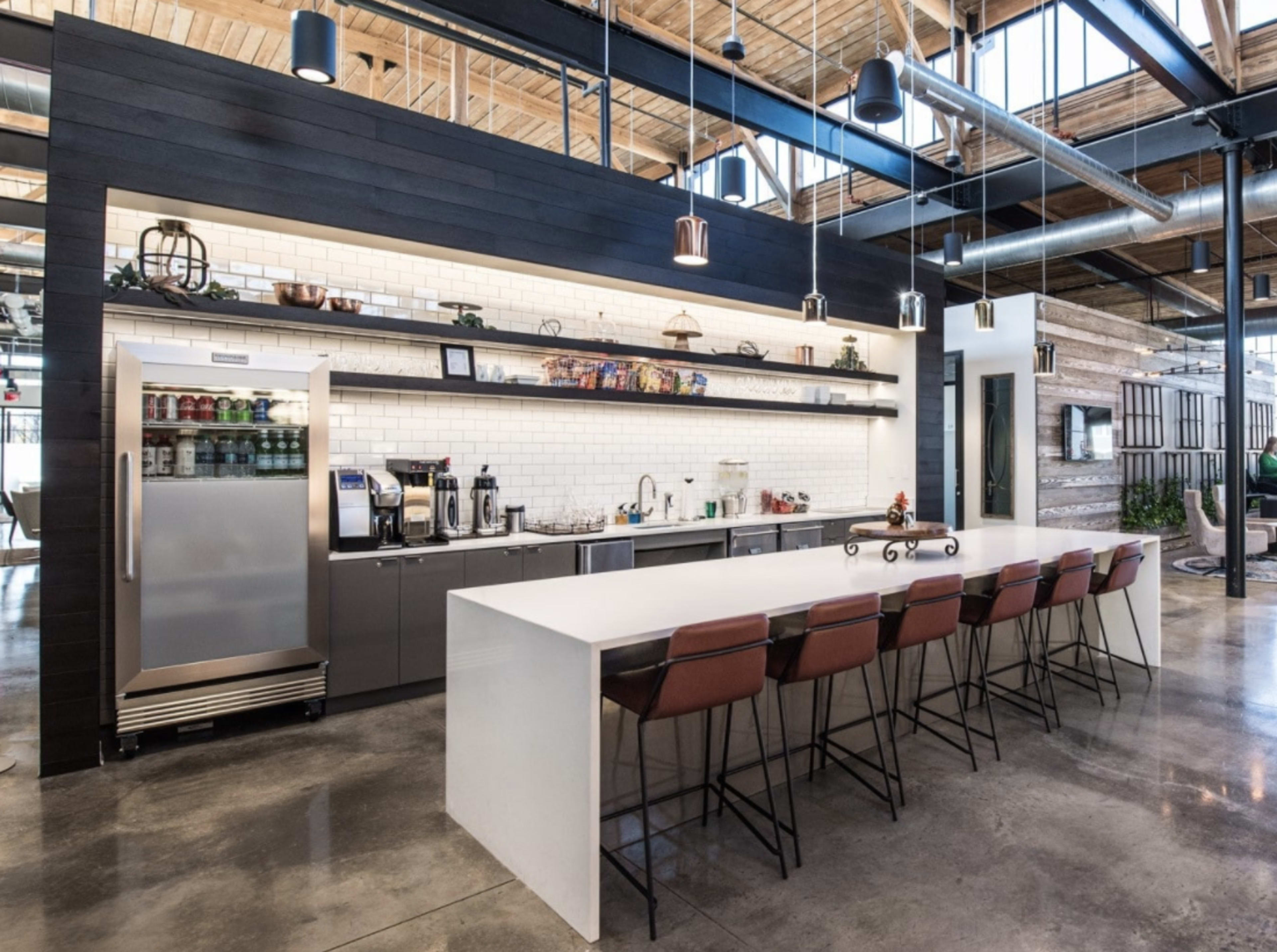 A modern kitchen area with a long white countertop, high-backed chairs, and stainless steel appliances against a backdrop of exposed wooden beams and polished concrete floors.