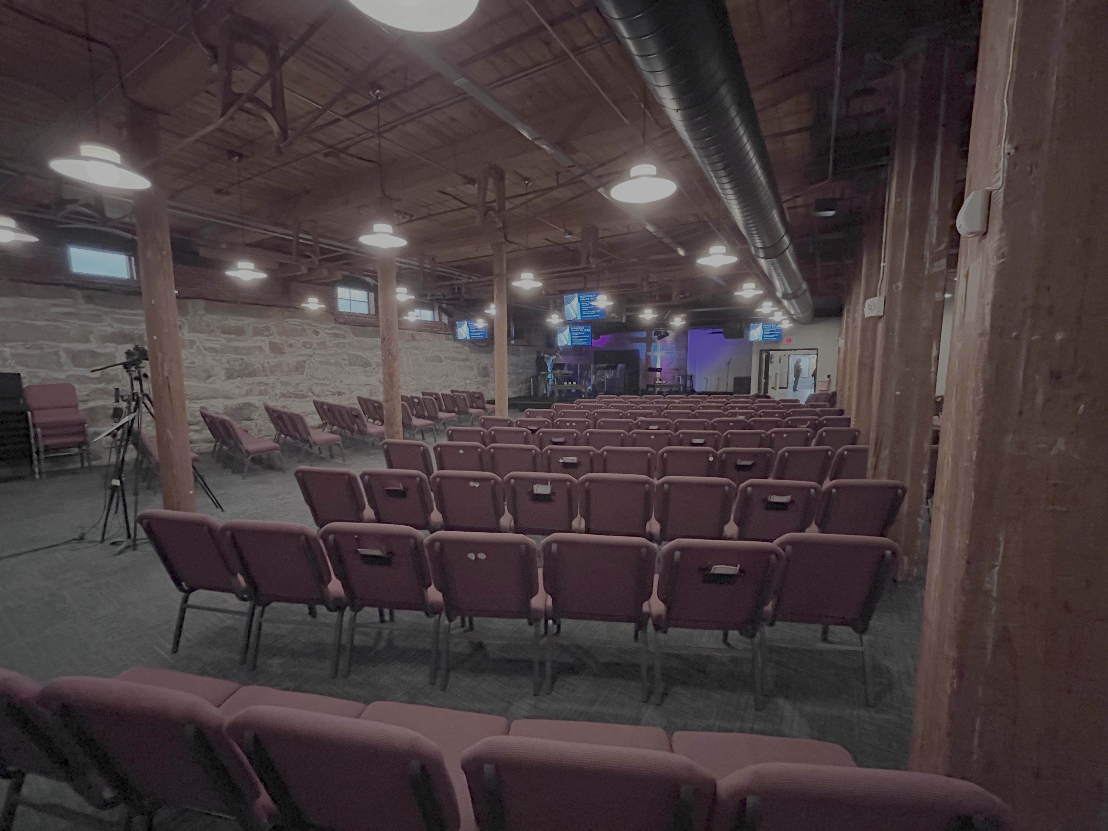 The image shows a spacious auditorium with rows of maroon chairs arranged facing a stage area, featuring exposed wooden beams and stone walls.