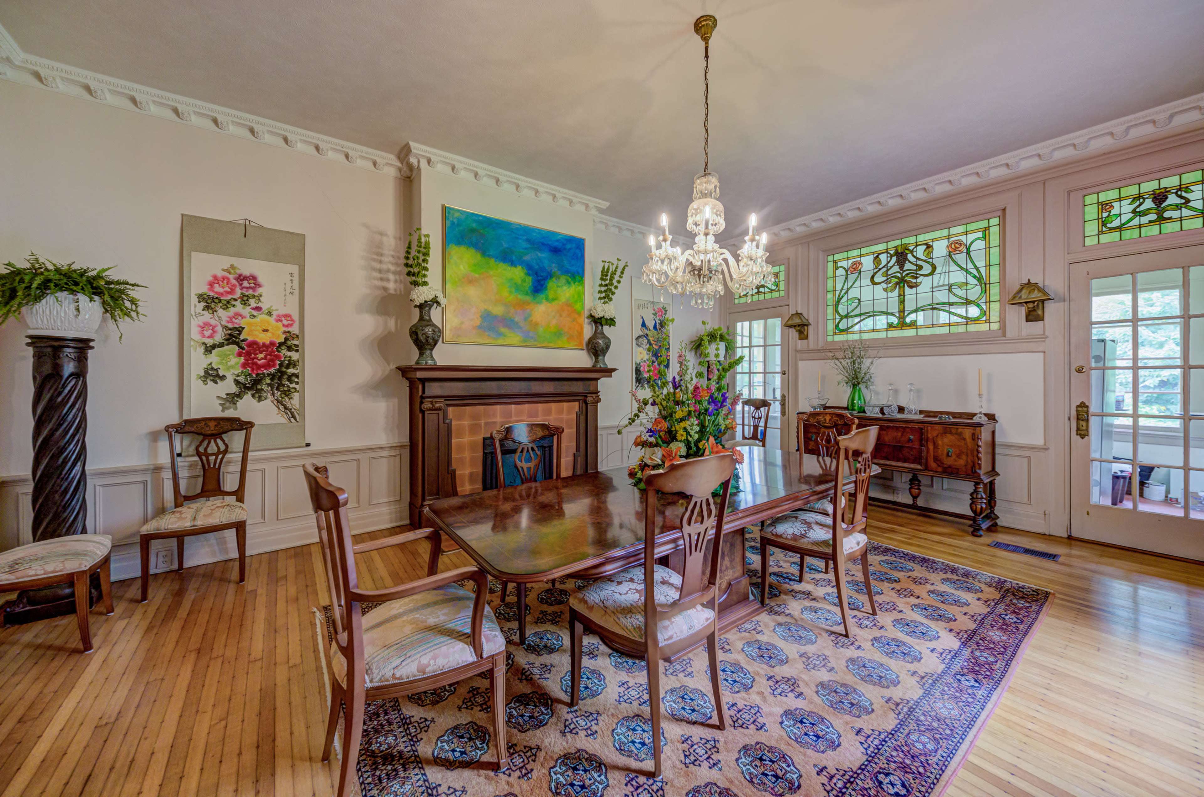 A dining room features a wooden table surrounded by chairs, a fireplace with a decorative mantle, and a chandelier above, accented by stained glass windows and wall art.