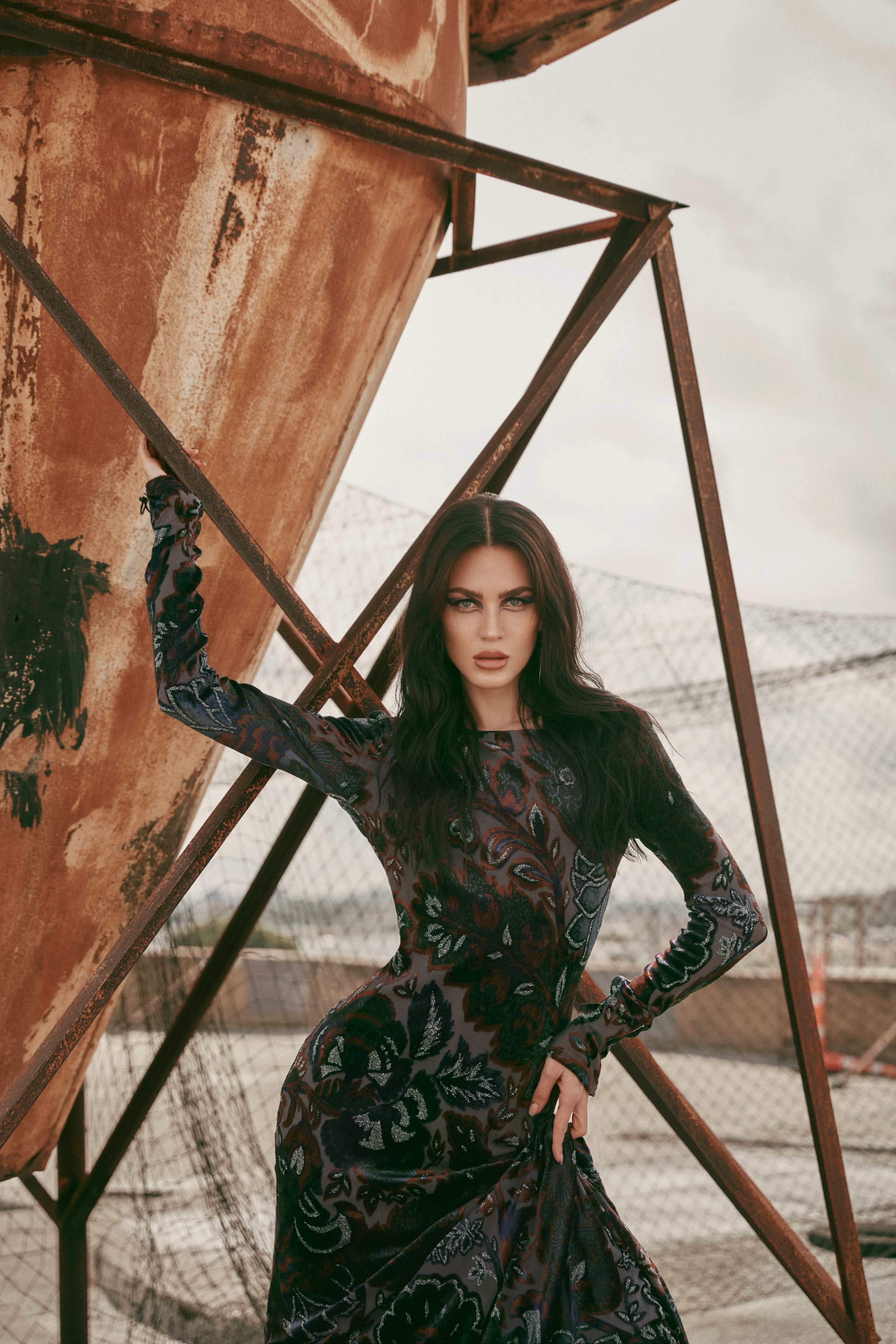 A woman in a patterned black dress poses against a rusted industrial structure.
