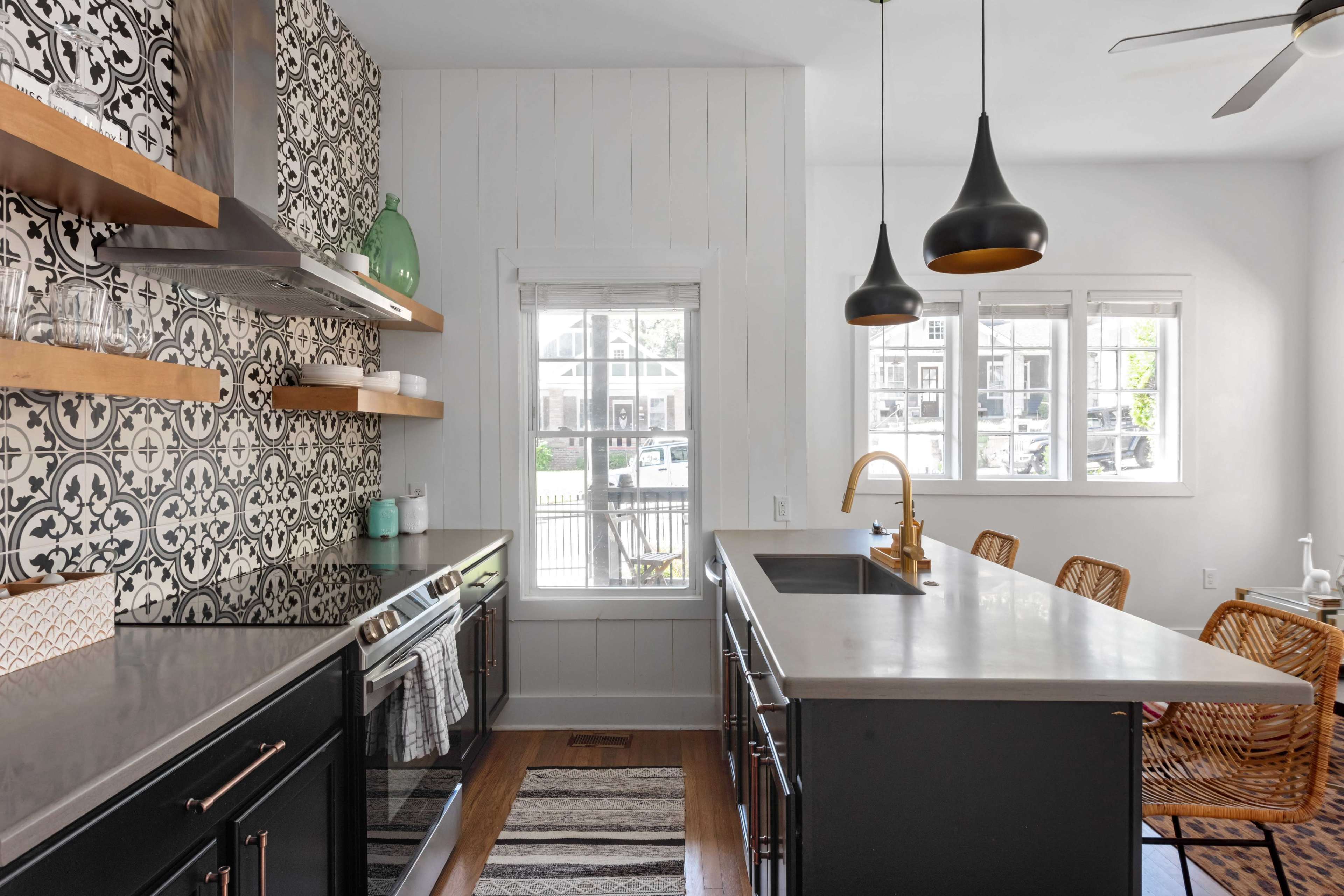 The image shows a modern kitchen with patterned tile backsplash, open shelving, and a central island equipped with pendant lights and rattan dining chairs.