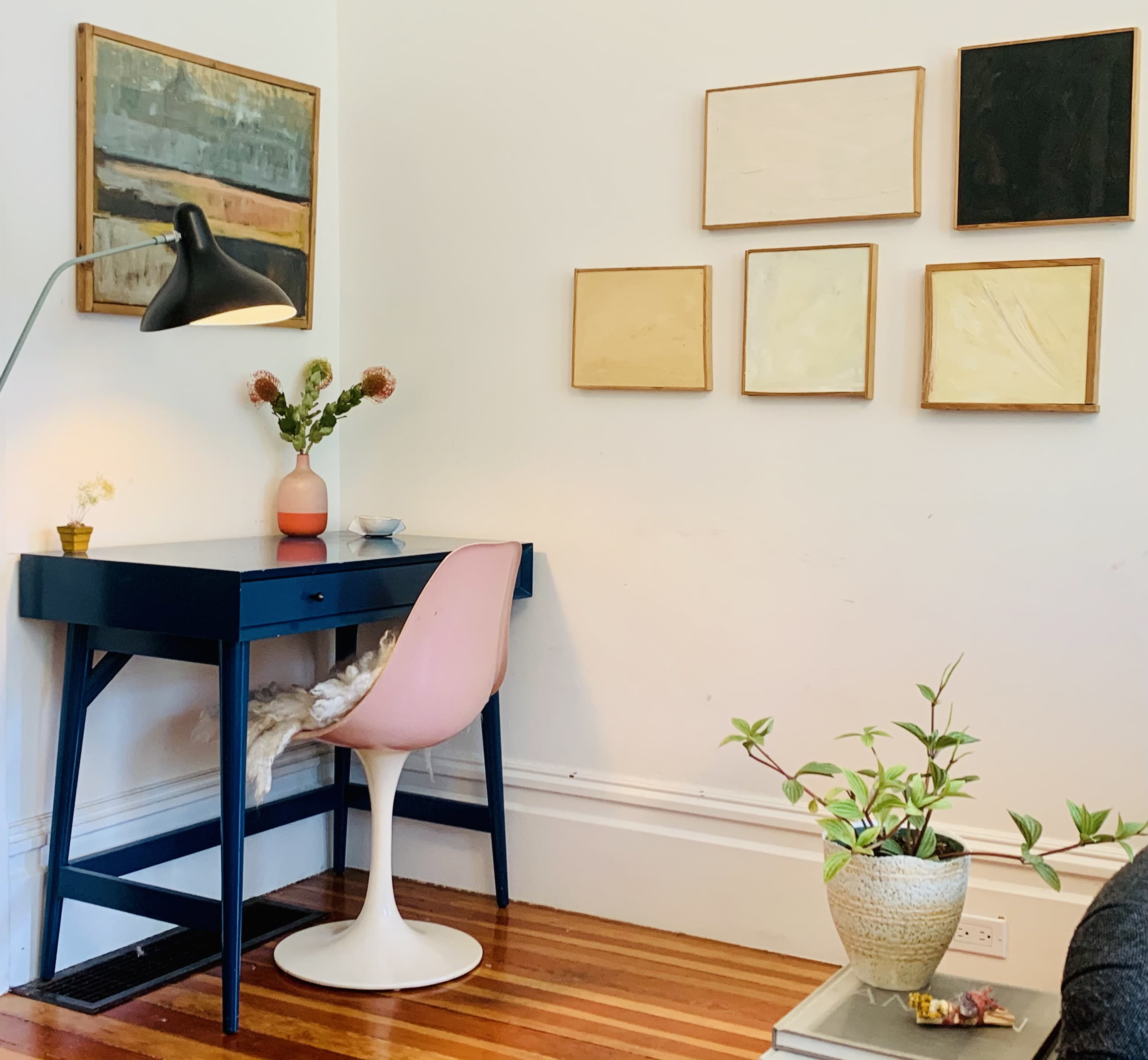 A small desk with a pink chair is positioned against a wall adorned with framed artwork, next to a lamp and a decorative plant.