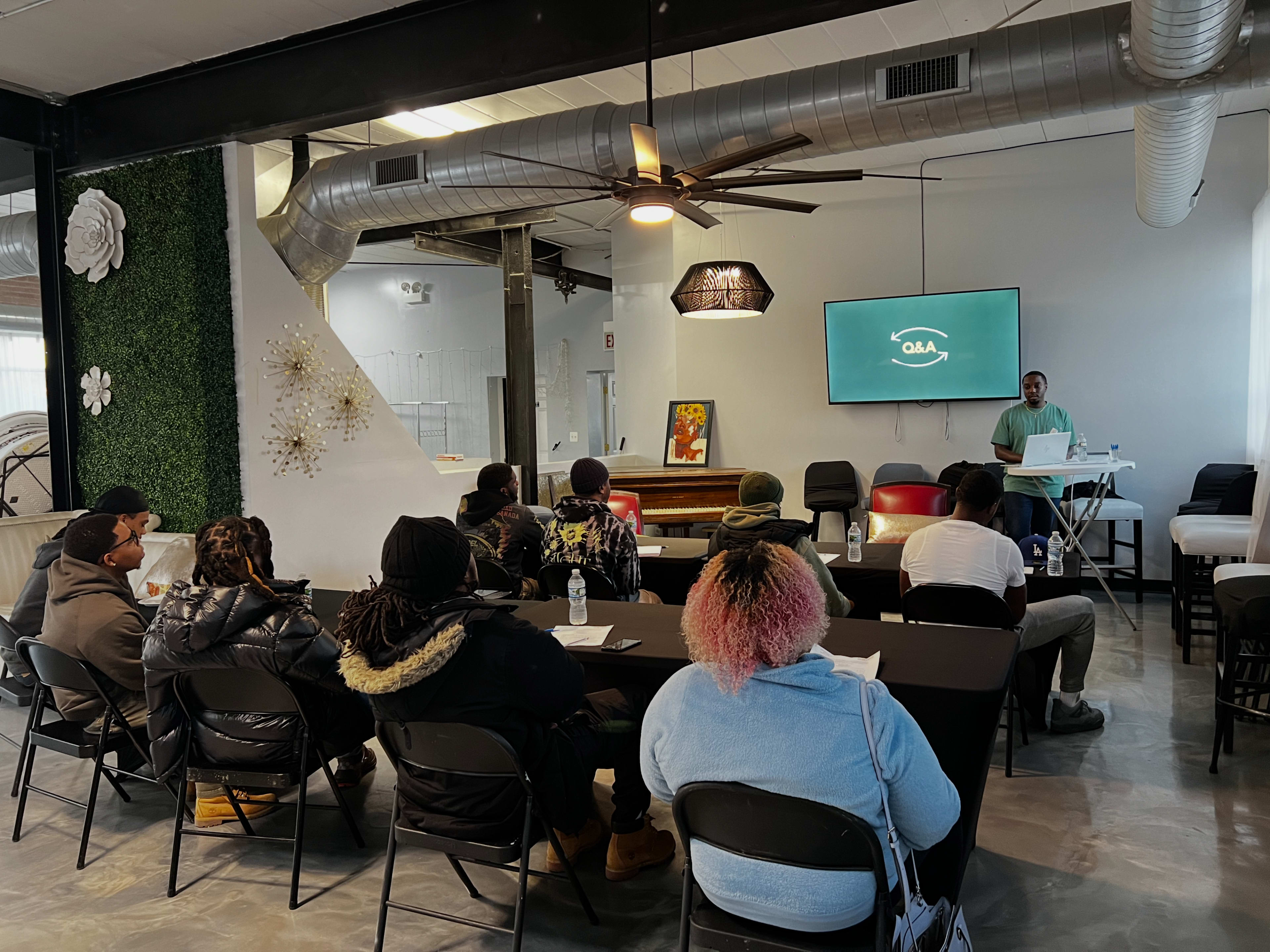 A group of people is seated at tables in a modern room, facing a presenter giving a talk in front of a screen.