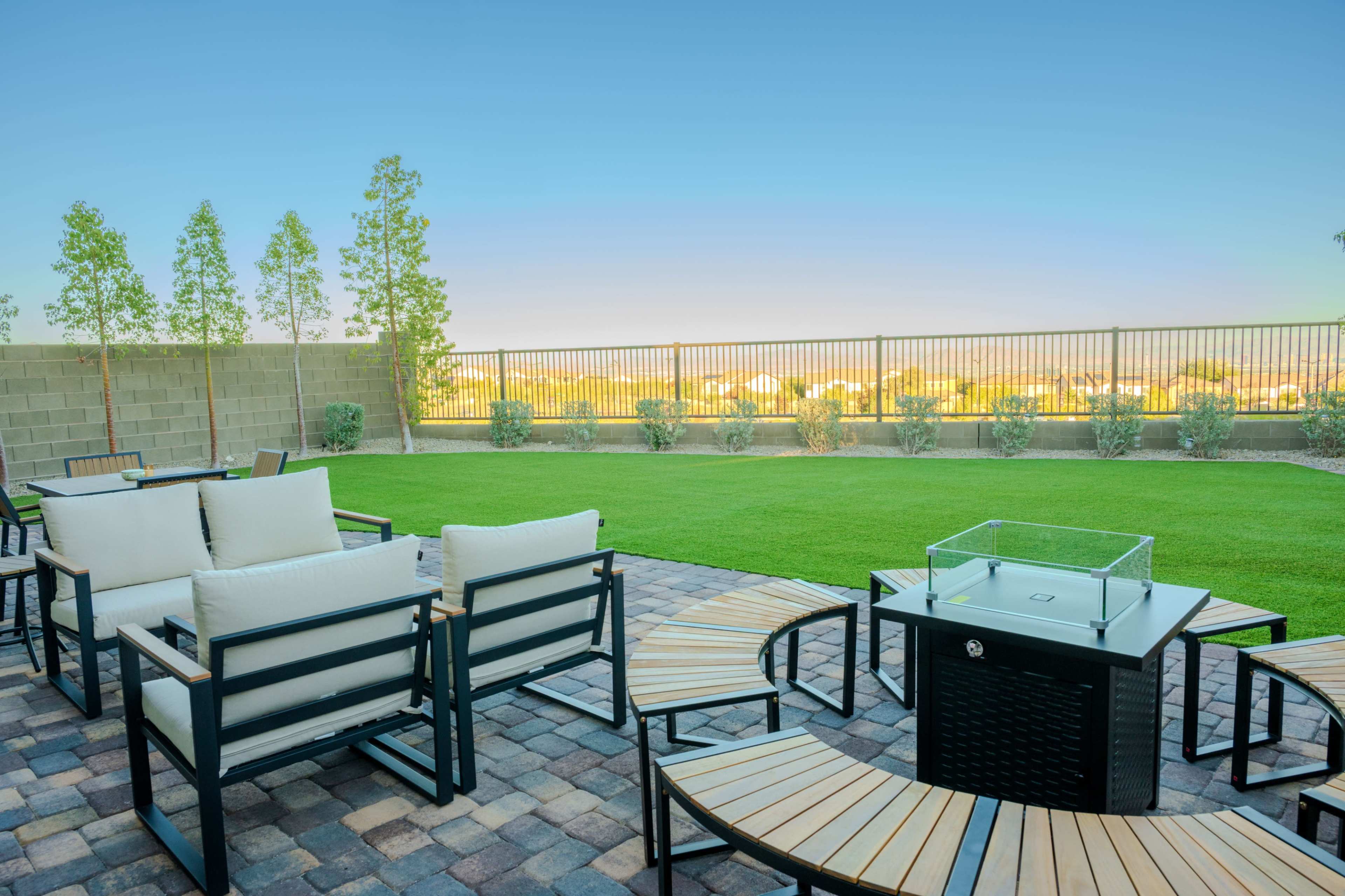 A patio features black-framed seating with light cushions arranged around a fire pit, set on a paved surface with a grassy area and a fence in the background.