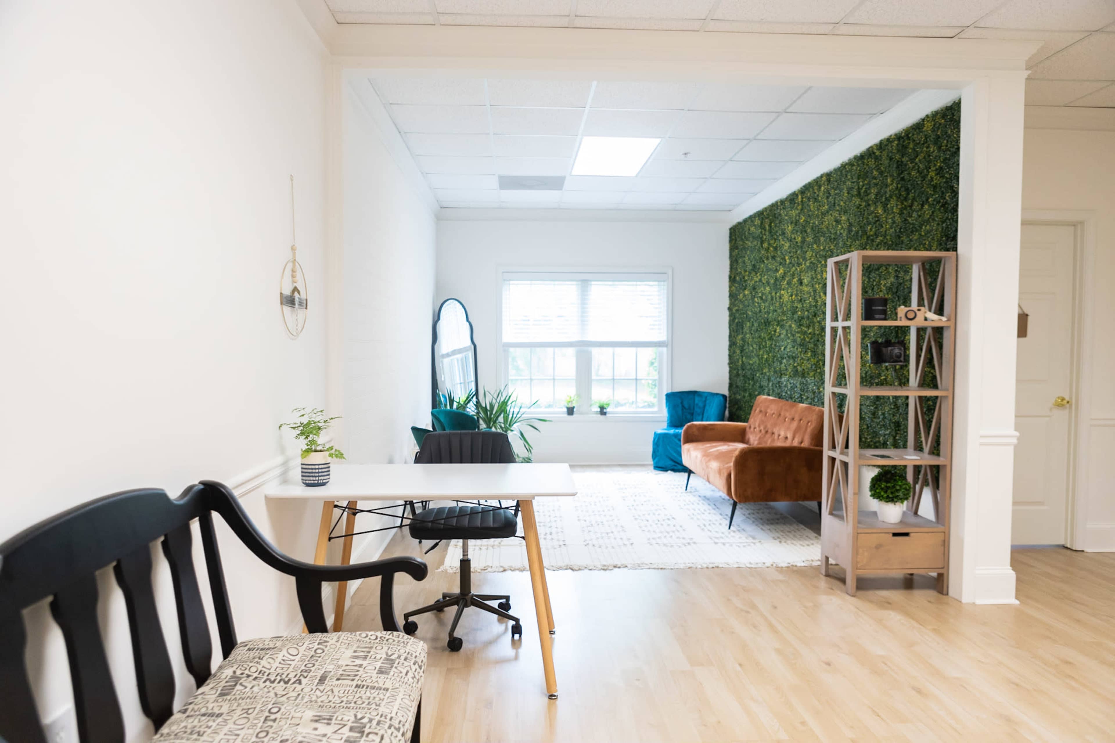 The image shows a modern office space featuring a desk and chair, a bench, two orange and teal couches, a bookshelf, and a green wall with plants.