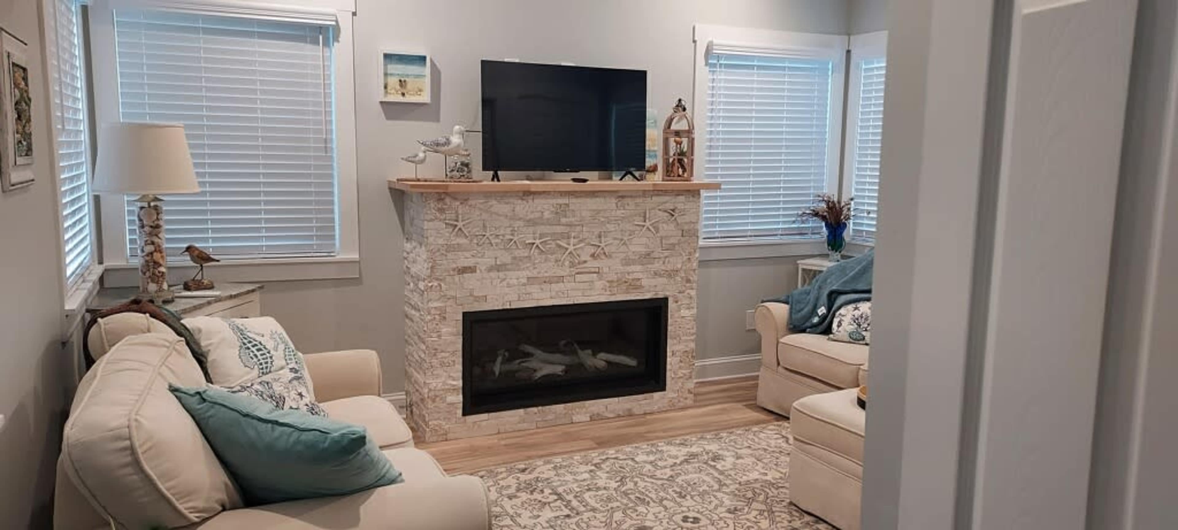 The living room features a stone fireplace below a television, flanked by two armchairs and a beige couch, with light-colored walls and window treatments allowing natural light.