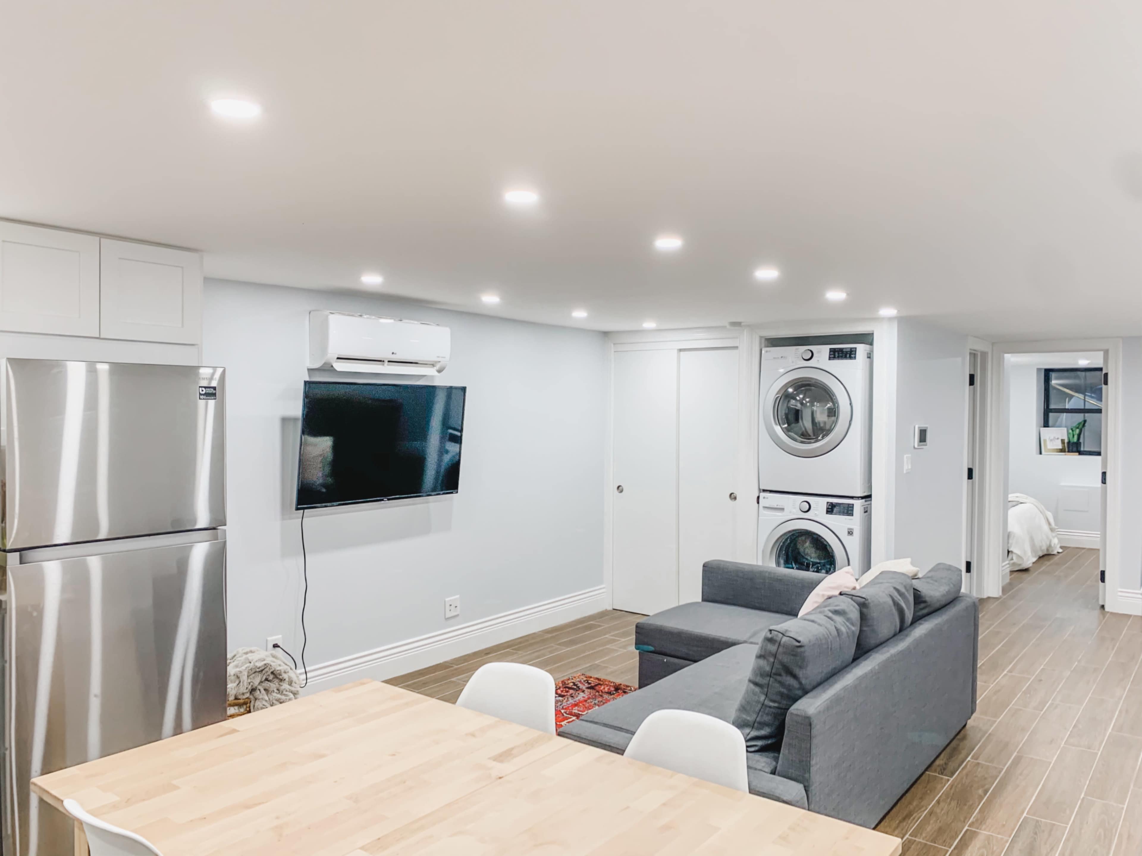 A modern, open-plan living space with a kitchen area, a sofa facing a wall-mounted television, a washer and dryer stacked next to a door, and a dining table nearby.