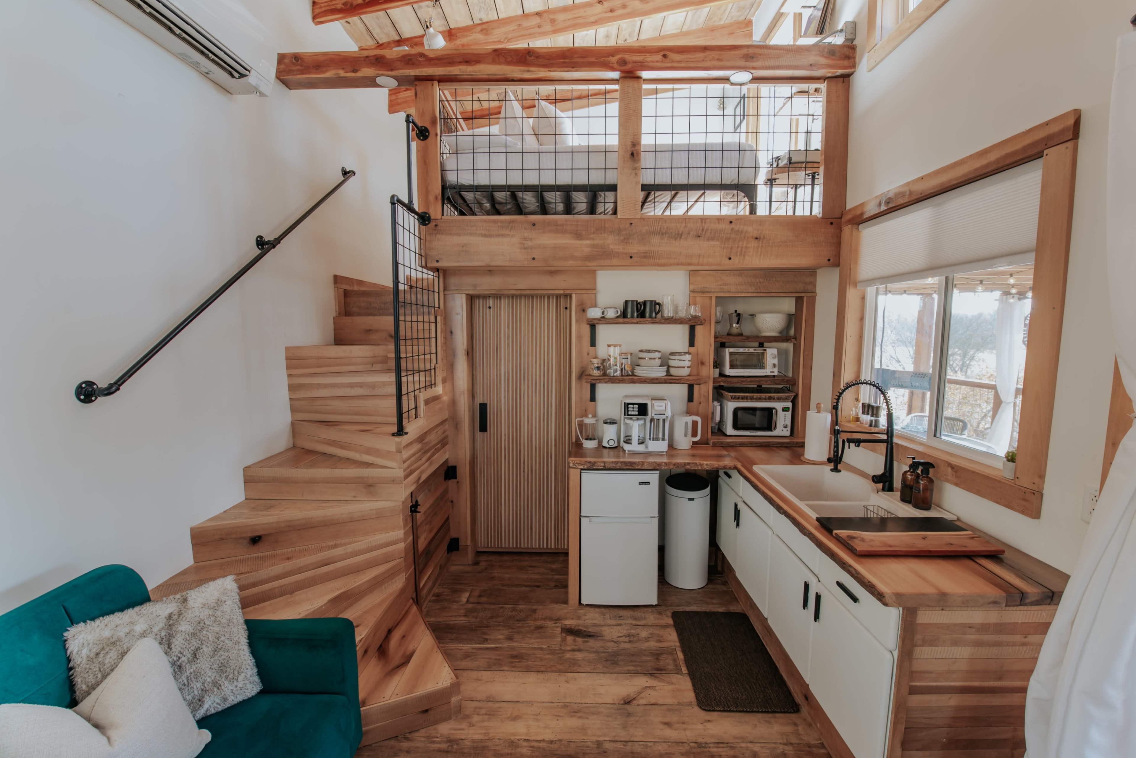 A cozy, wooden interior of a small house with a staircase leading to an upper loft and a kitchen area equipped with appliances.