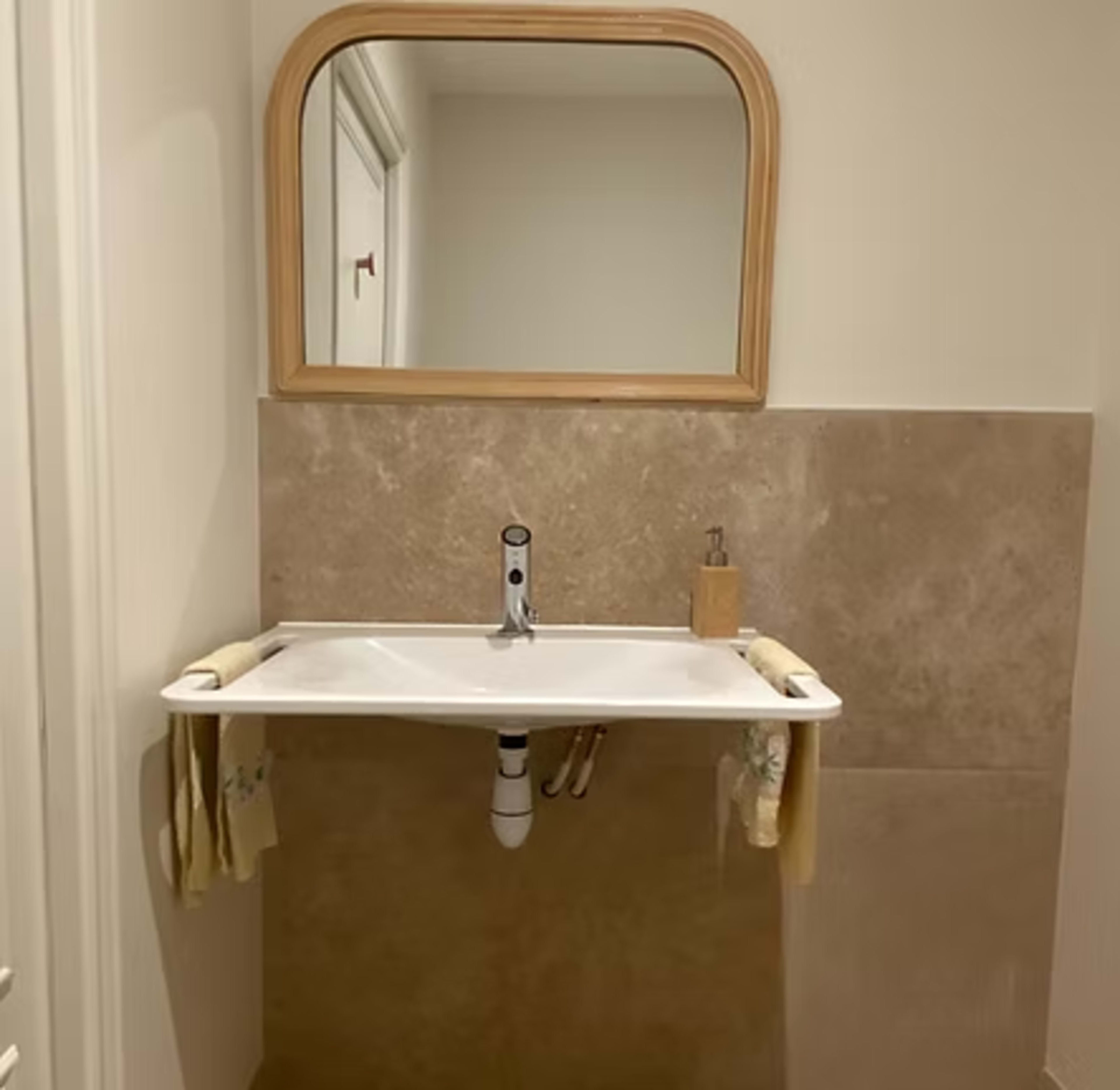 The image shows a minimalist bathroom with a wall-mounted sink, a wooden-framed mirror, and beige tiled walls.