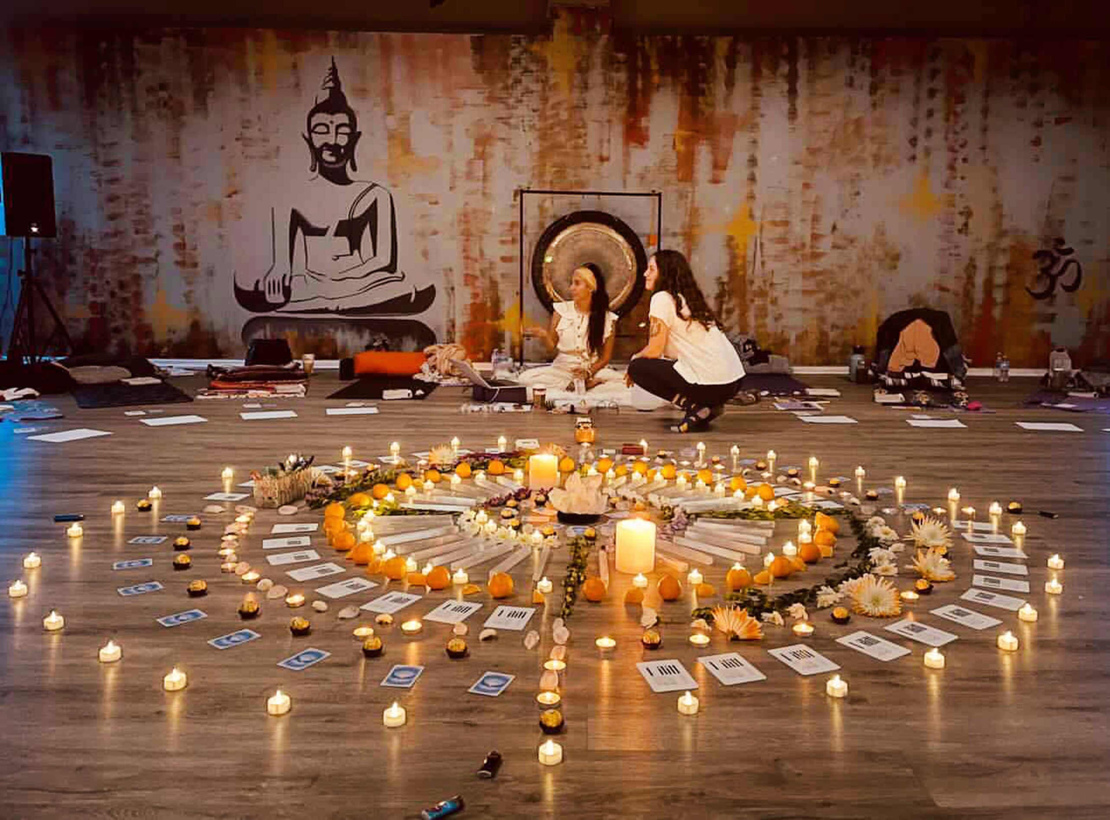 Two women sitting in a circular arrangement of candles, flowers, and cards in a dimly lit space decorated with a Buddha mural.