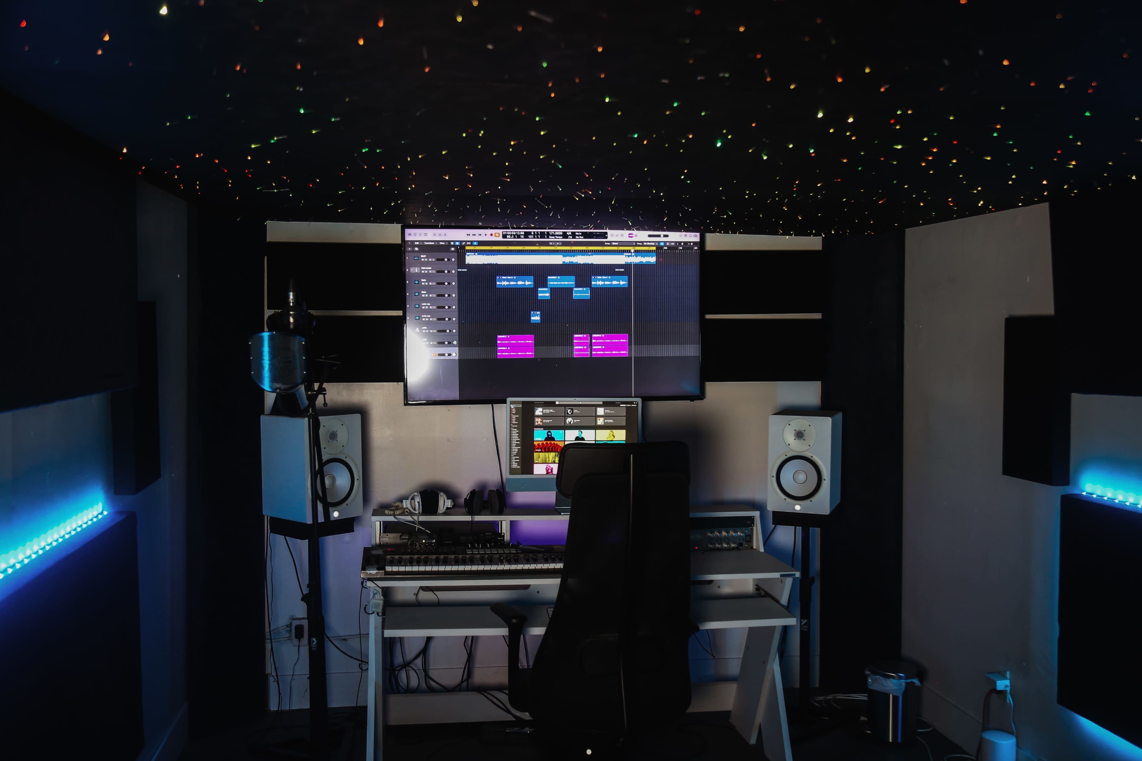 A modern music production studio features a desk with recording equipment, monitors, and a large screen displaying a software interface, all under a ceiling adorned with decorative lights.