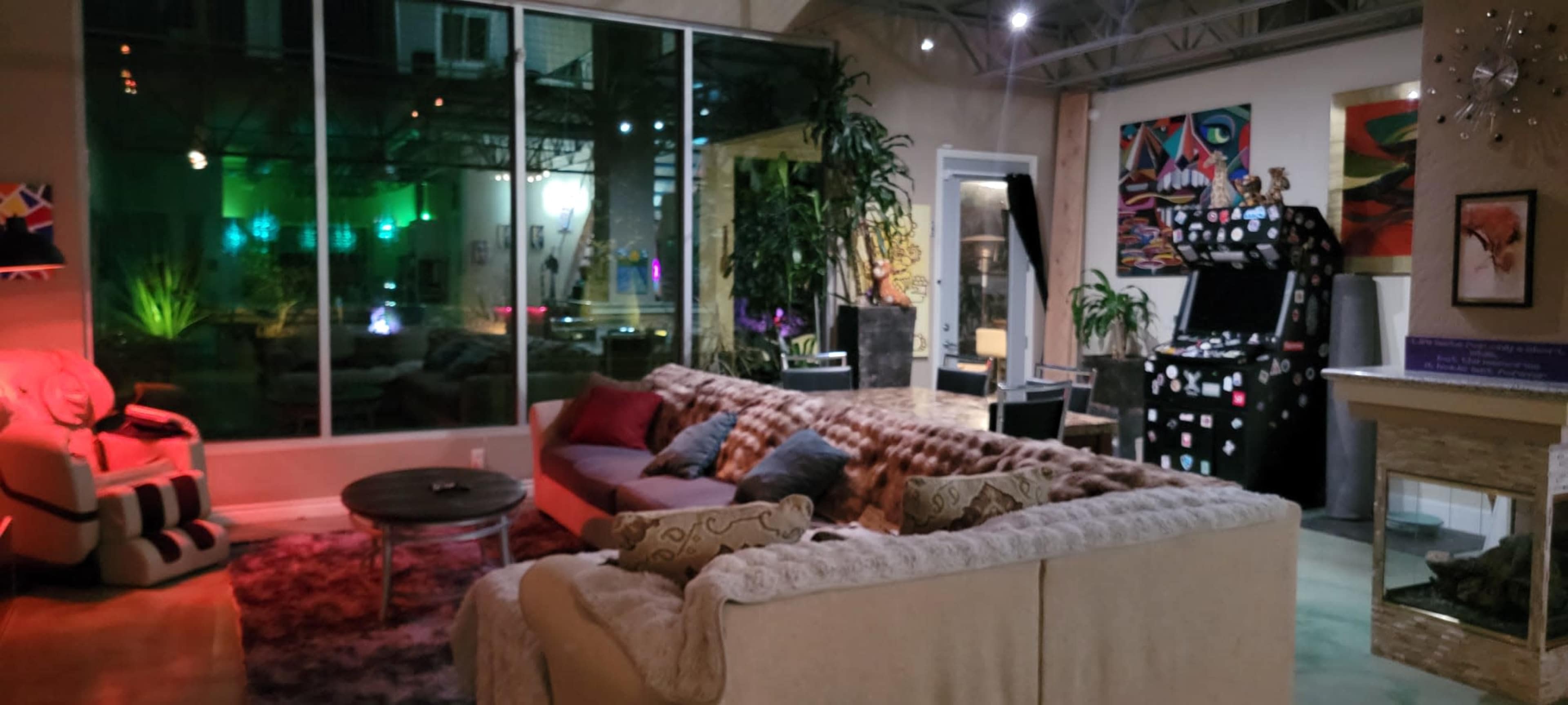 The image shows a modern living room with a large sectional sofa, a circular coffee table, and a view of a brightly lit outdoor space through large windows.