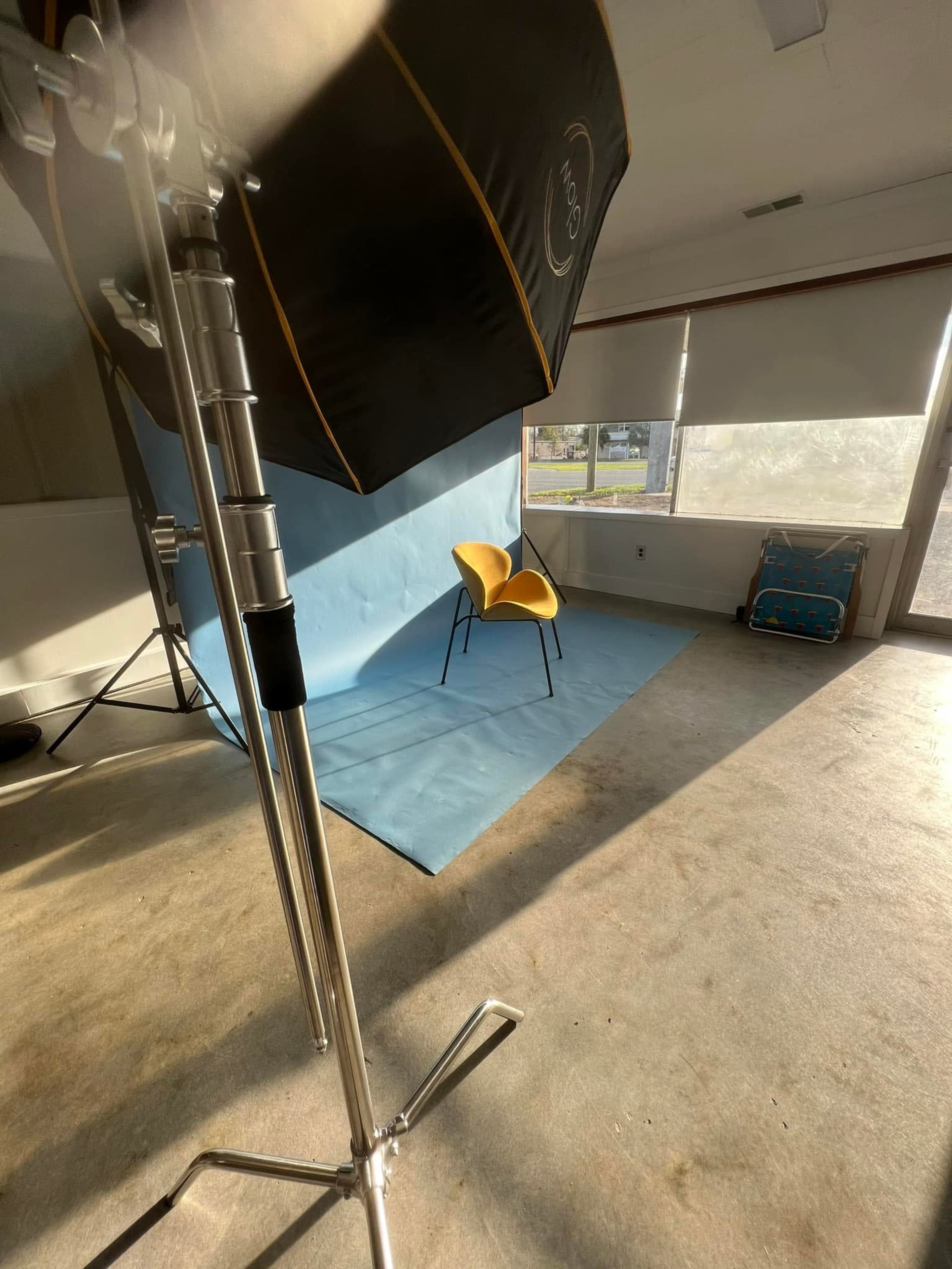 A studio setup features a yellow chair on a blue backdrop with softbox lighting and large windows providing natural light.