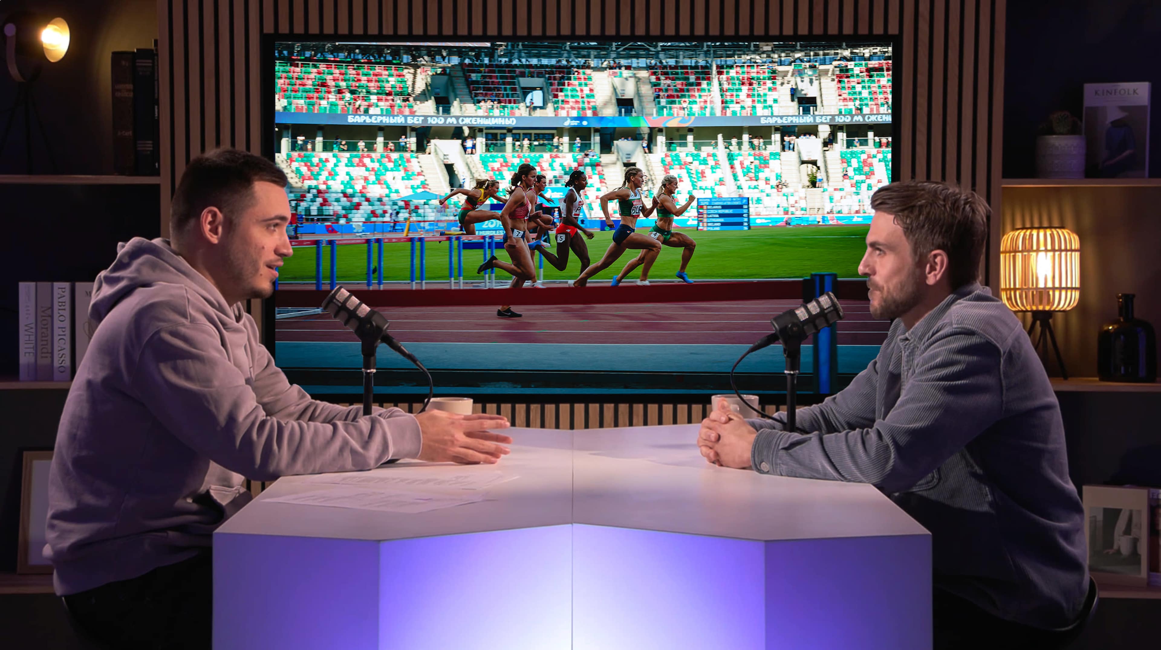 Two men are seated at a table with microphones, discussing in front of a large screen displaying athletes competing in a hurdle race.