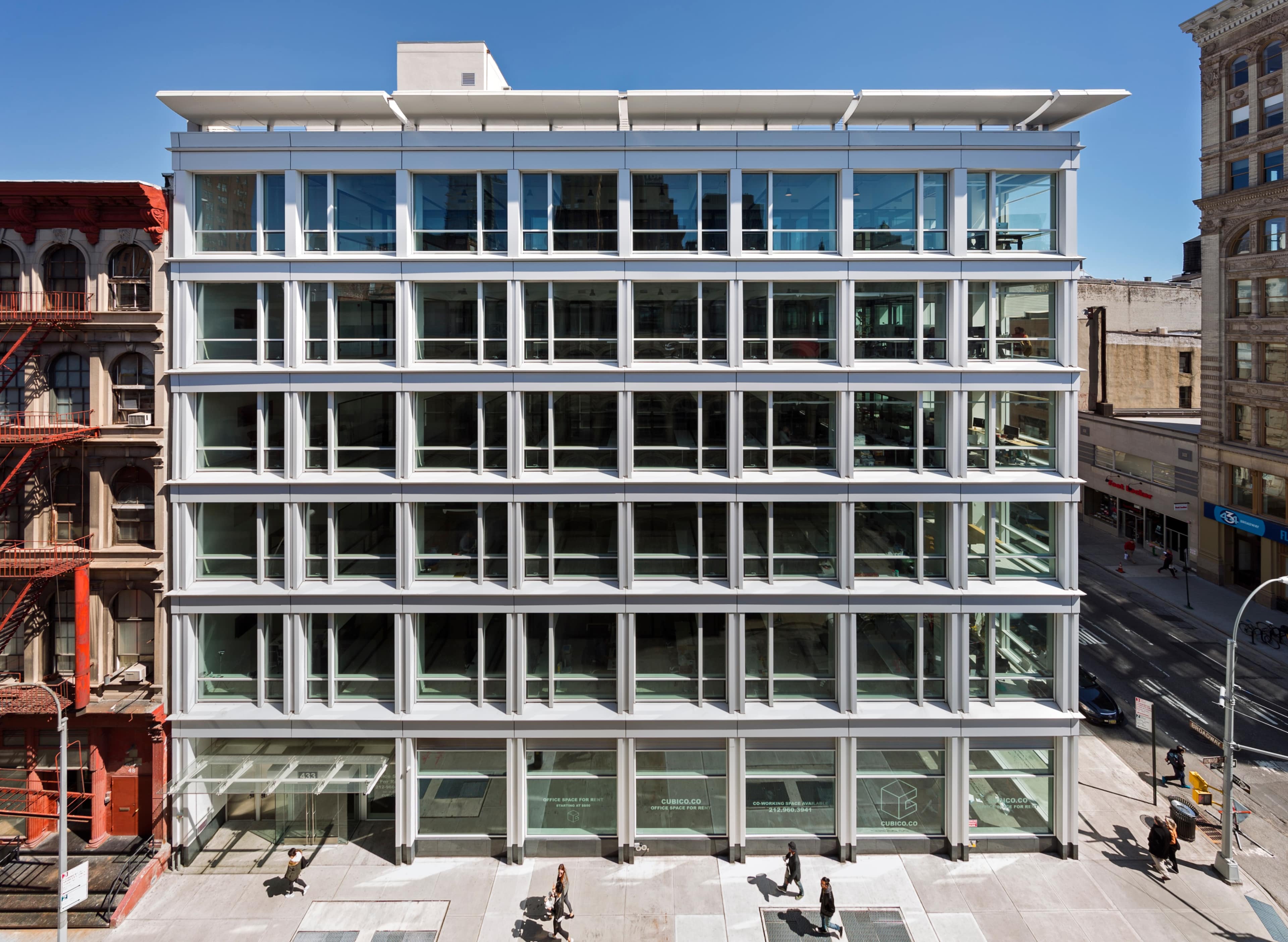 A modern, multi-story glass building features a grid of large windows facing a city street, with pedestrians walking by on the ground level.