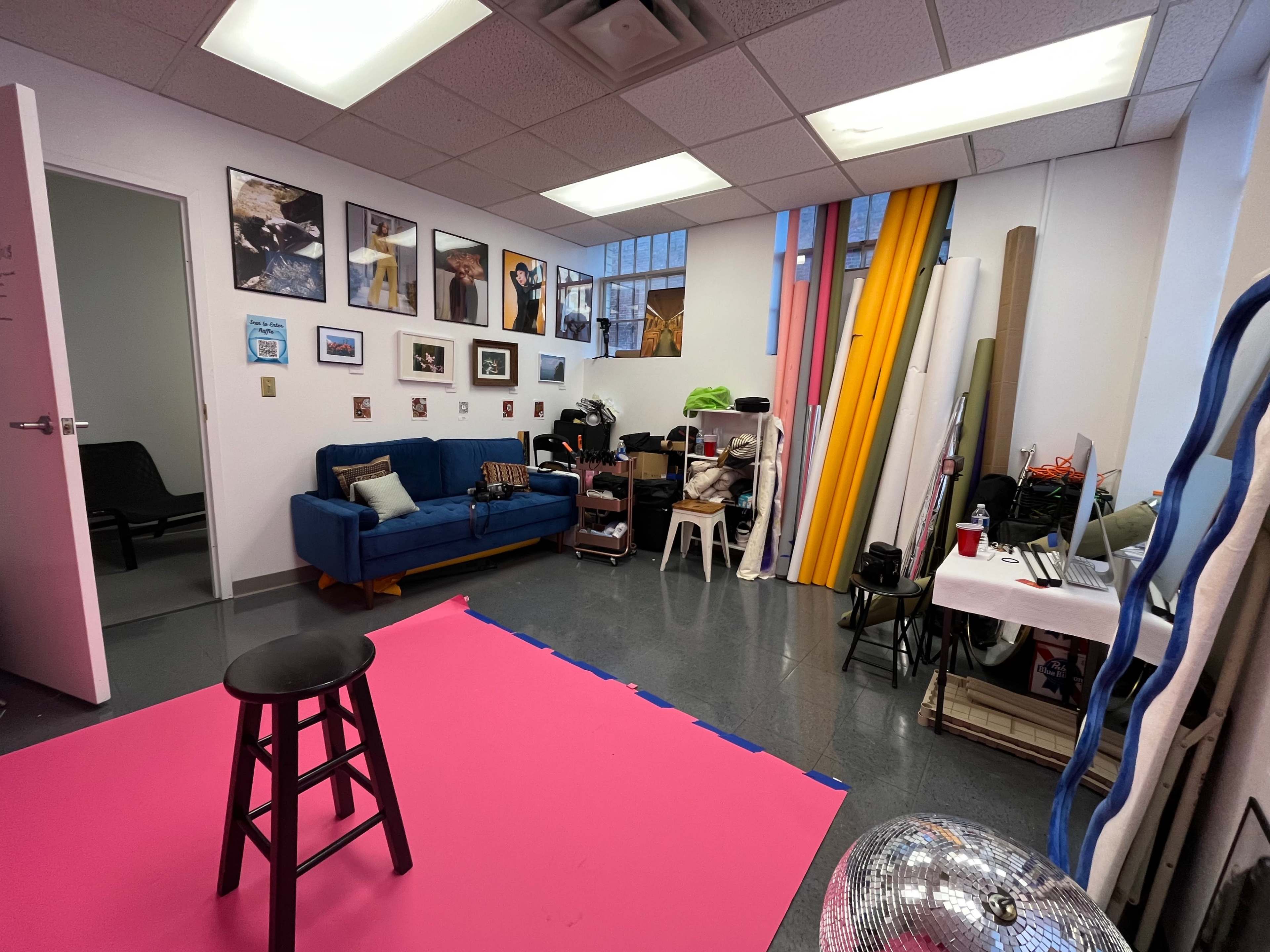 A studio space with a blue sofa, various framed pictures on the walls, a pink floor mat, and equipment scattered around, including a stool and rolls of colored backdrop paper.