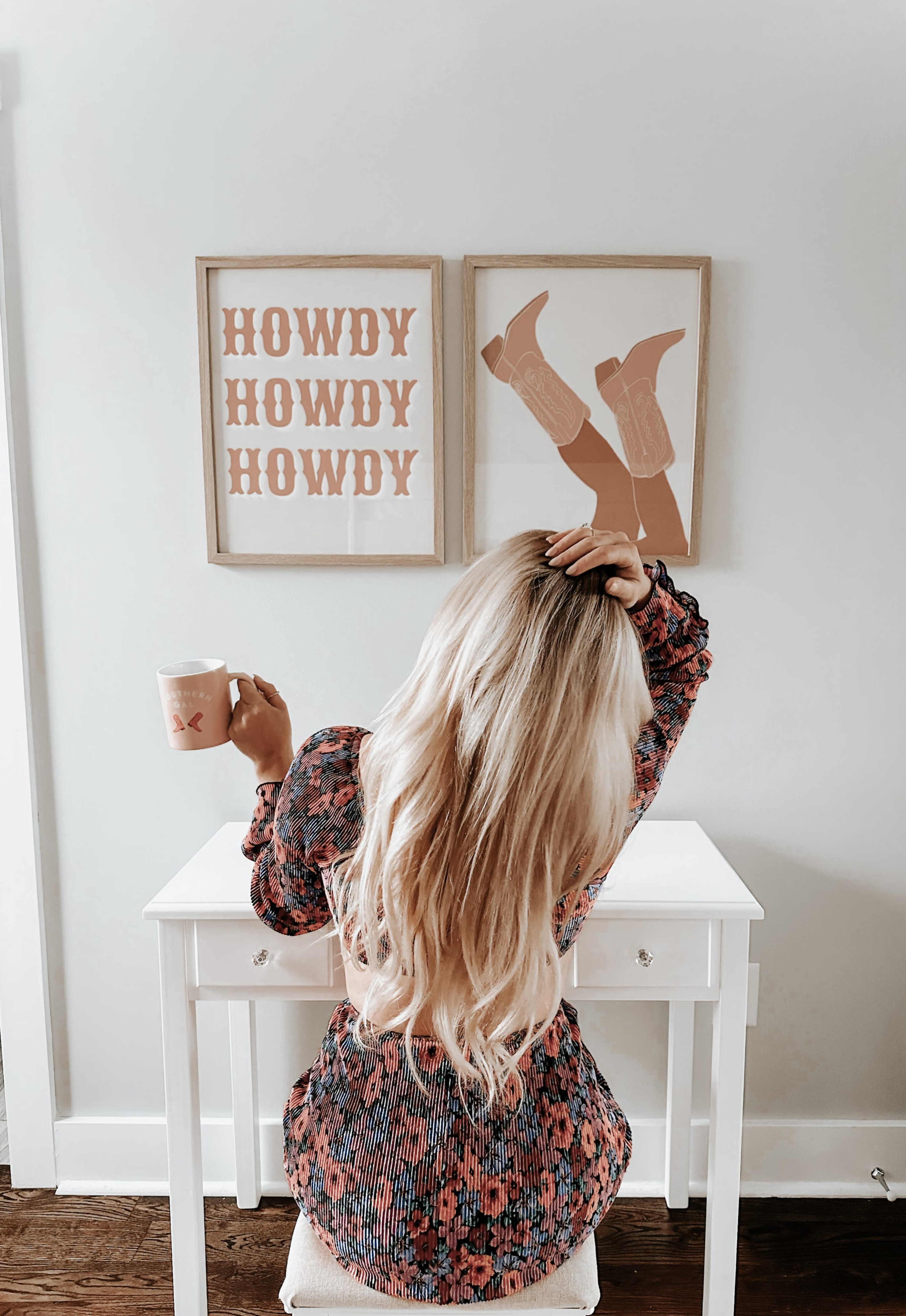 A woman sits at a small white desk, holding a mug, with her back to the camera, and two framed art pieces on the wall that say "HOWDY" in bold text.