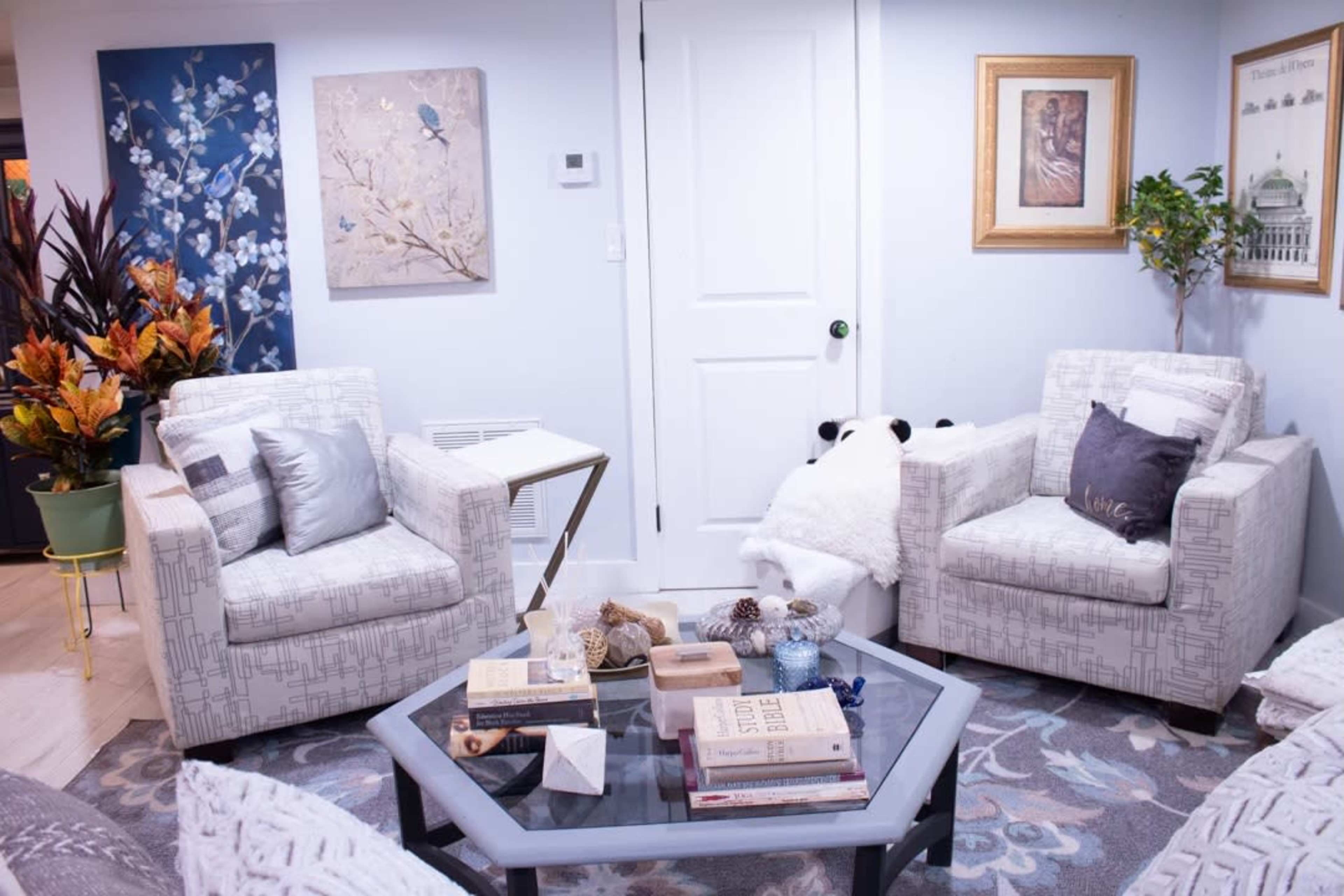 The image shows a cozy living room with two armchairs facing a coffee table, surrounded by decorative plants and wall art.