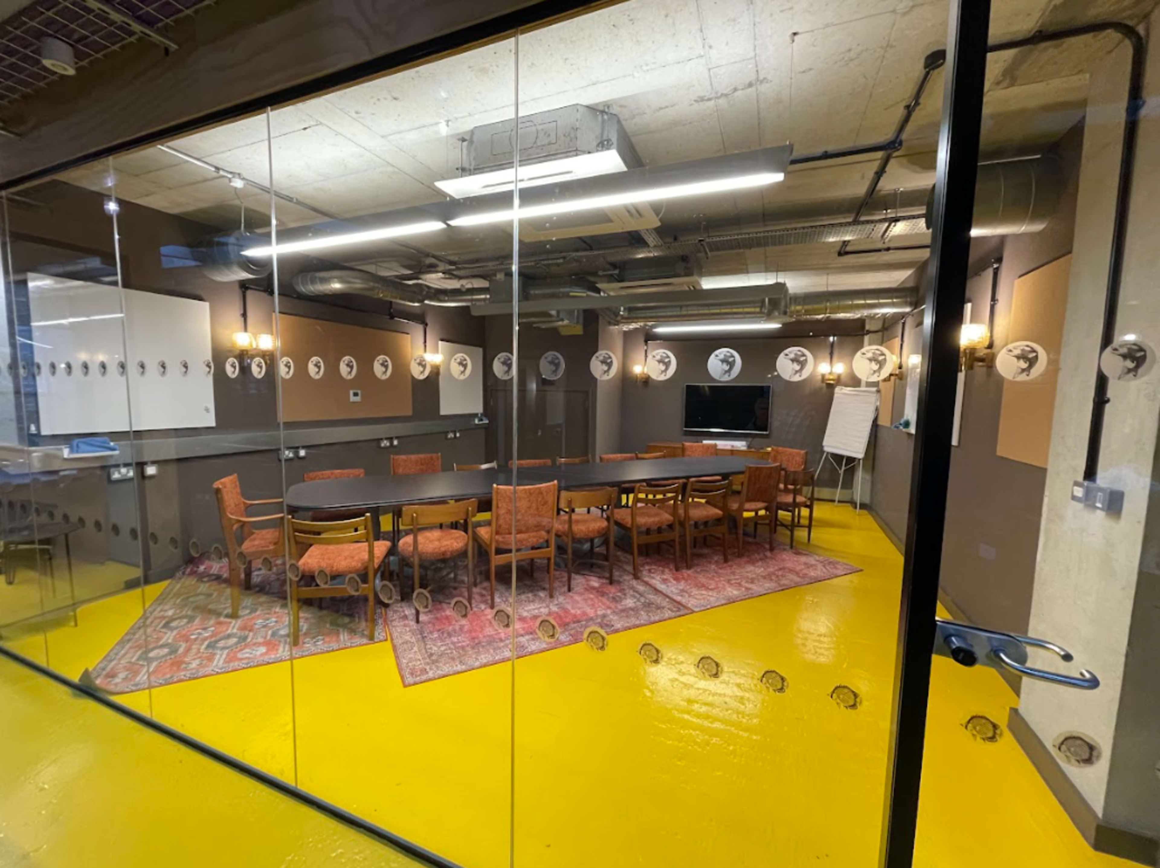 A conference room furnished with a long table surrounded by chairs, featuring a carpet and equipped with a projector screen, is enclosed by glass walls.