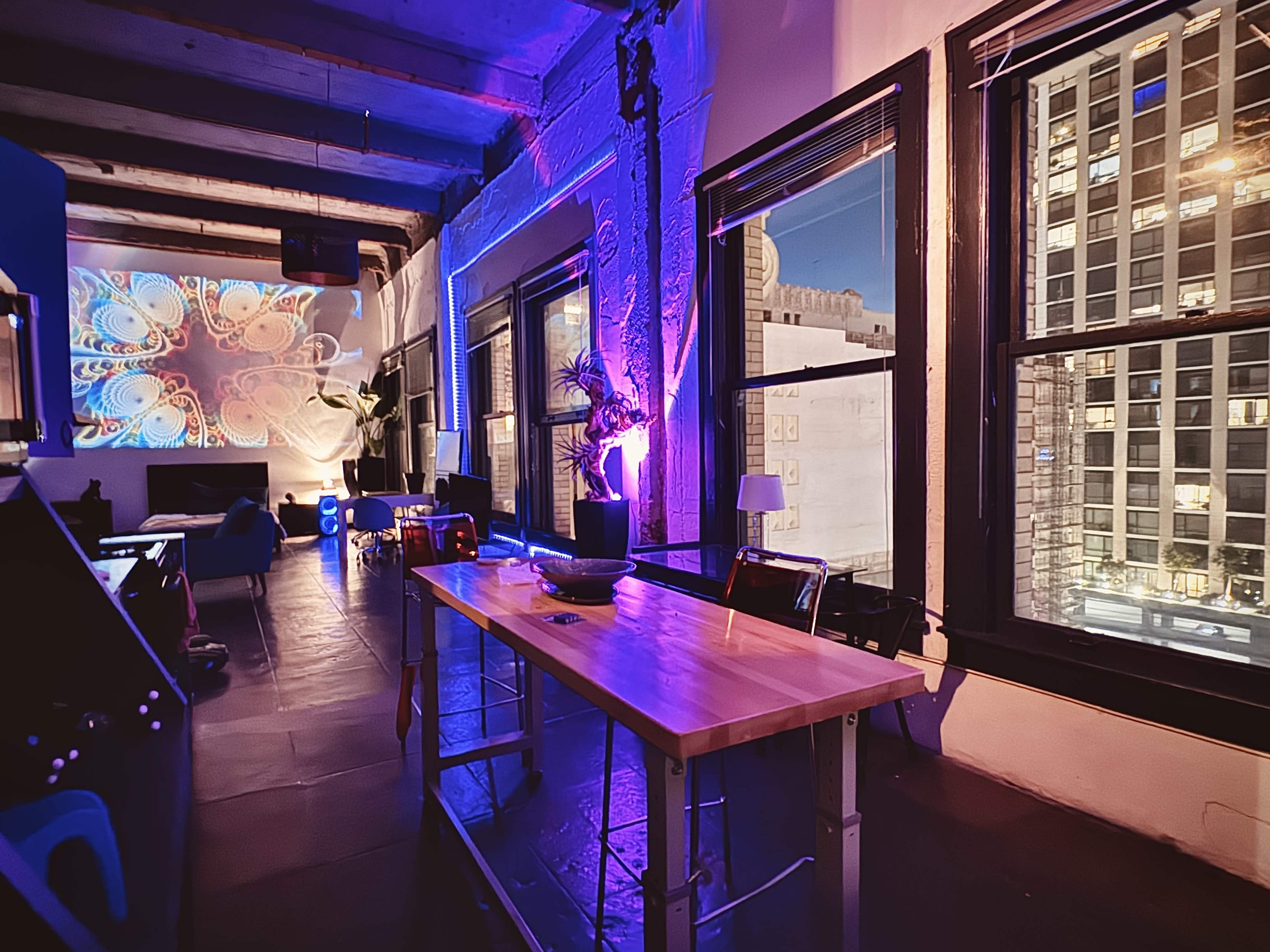 A modern loft space with a wooden table, colorful wall projections, and large windows revealing a nighttime city view.