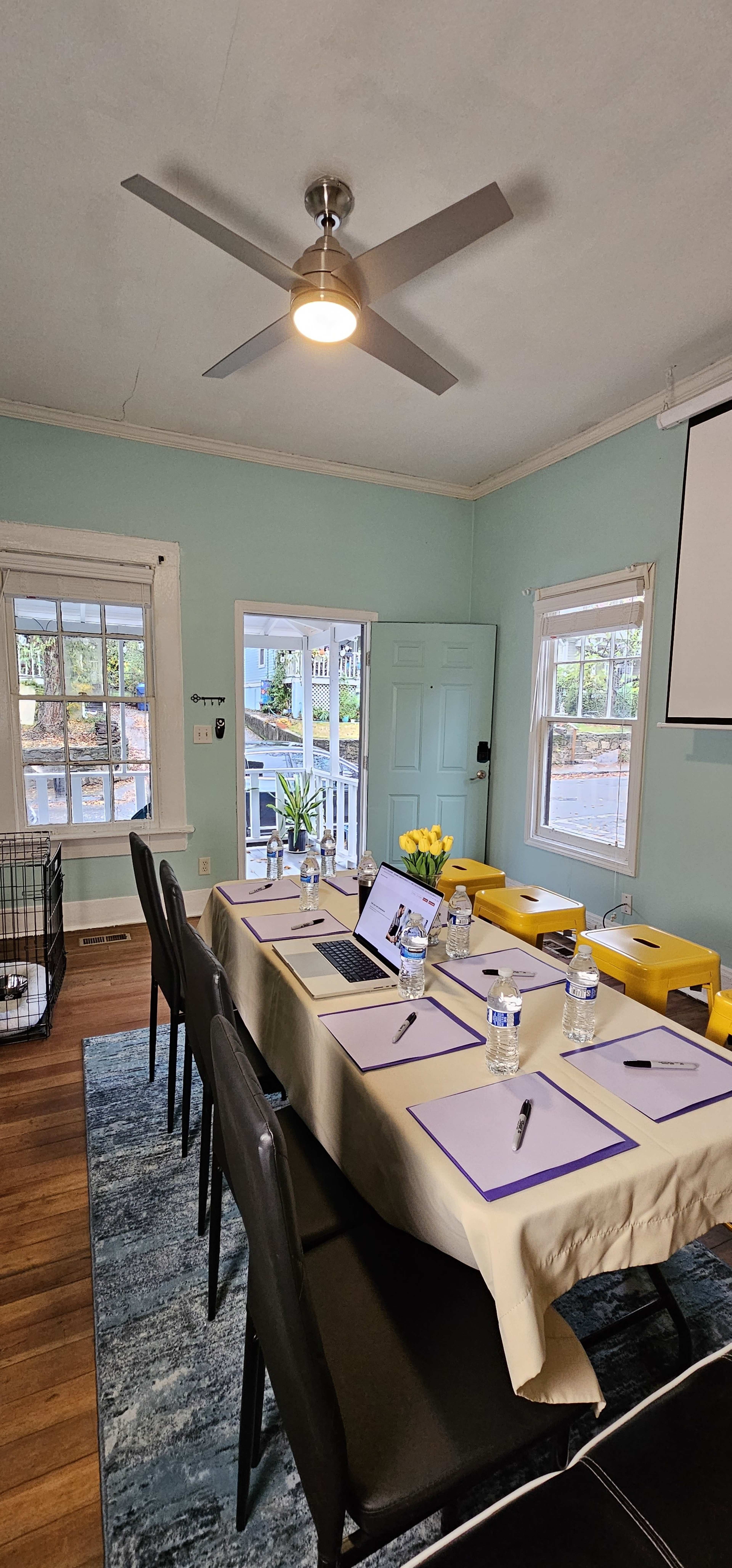 A meeting room is set up with a long table covered in papers, water bottles, and a laptop, flanked by chairs and a projector screen.