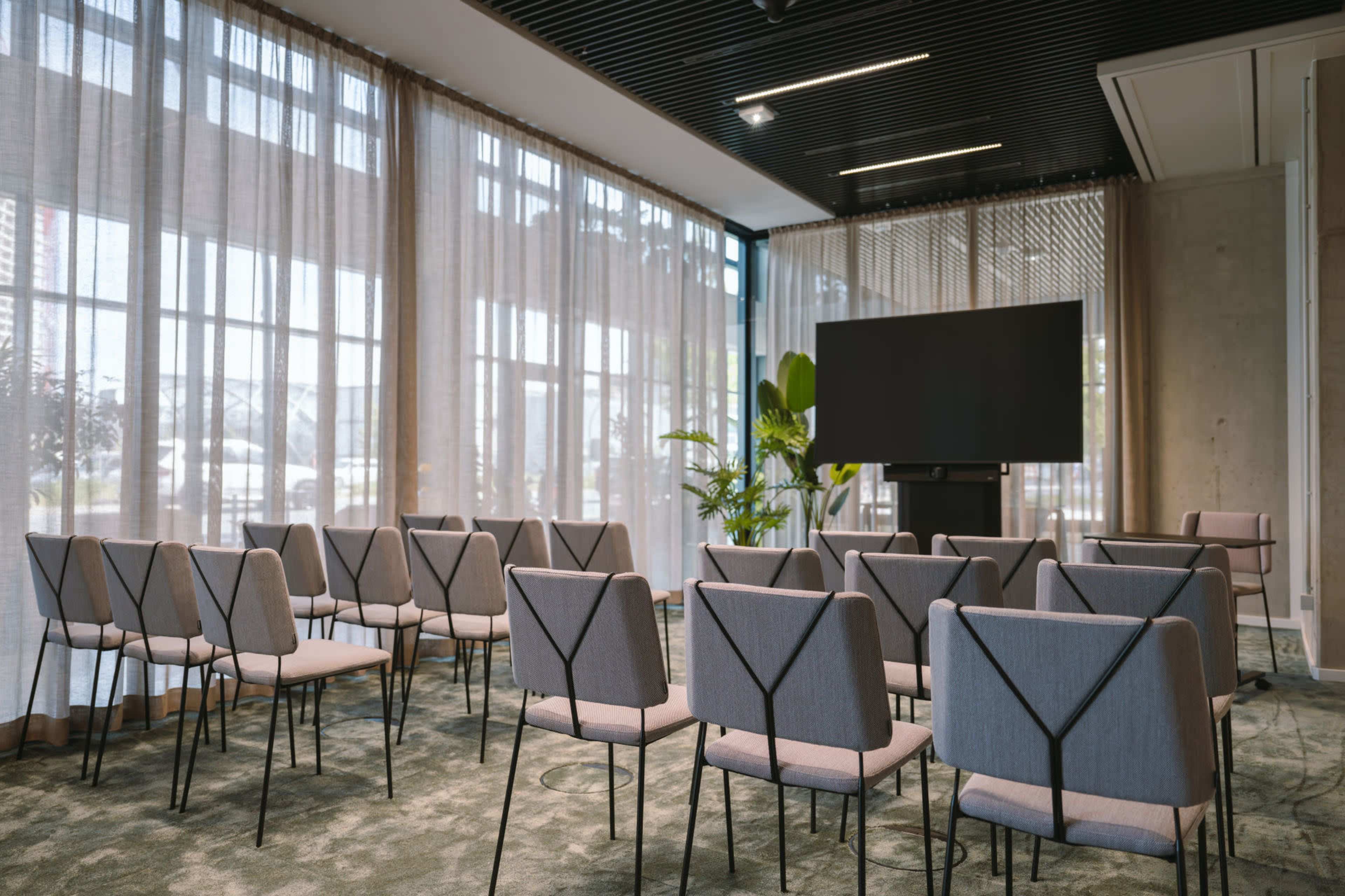 A conference room is set up with rows of chairs facing a large screen and large windows letting in natural light.