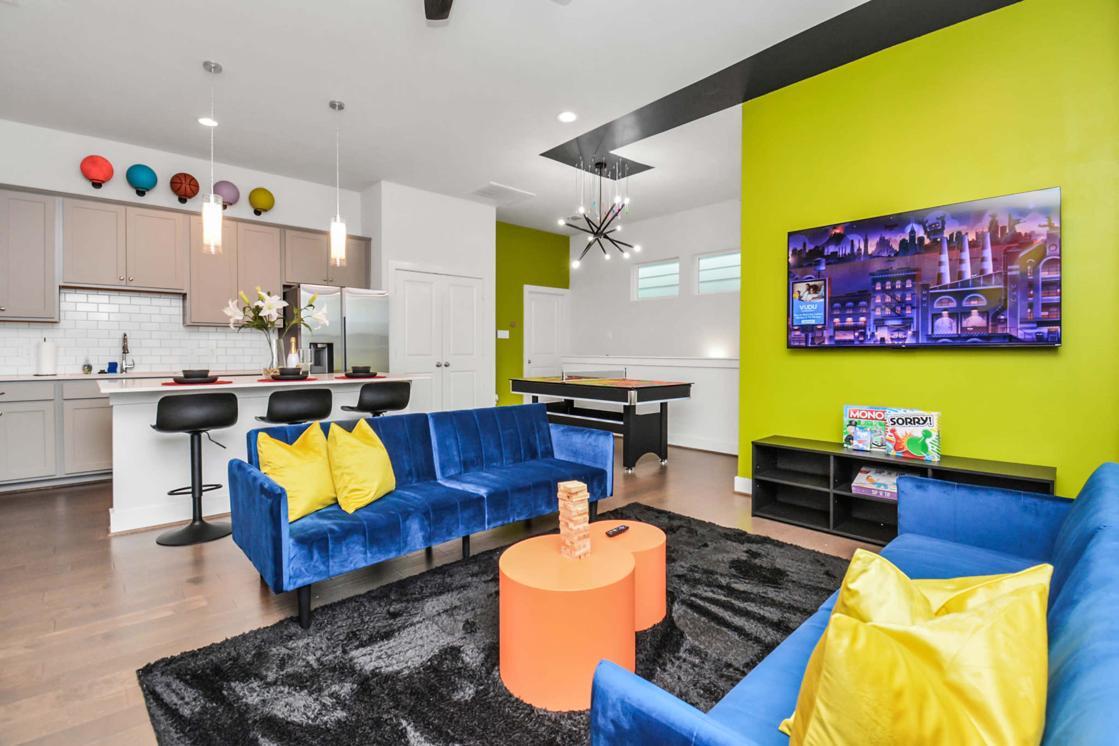 The room features a bright green accent wall, blue sofas arranged around an orange coffee table, and a modern kitchen visible in the background.