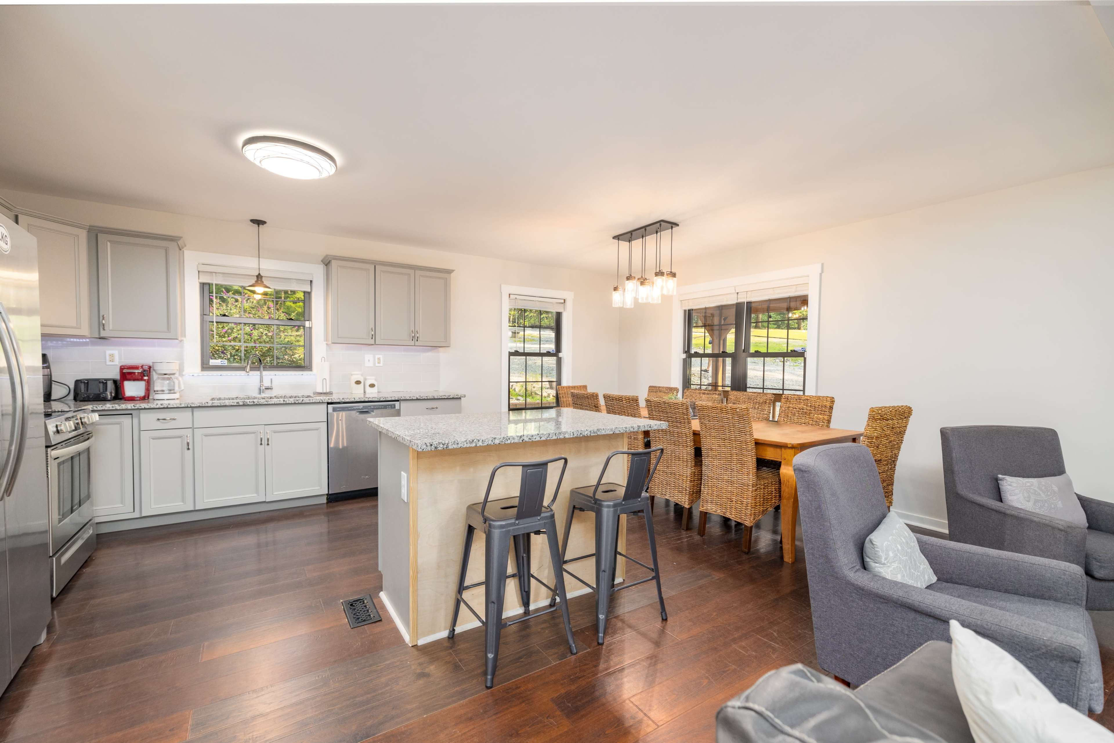 The image shows a modern kitchen and dining area with gray cabinets, a central island with bar stools, and a wooden dining table surrounded by woven chairs.