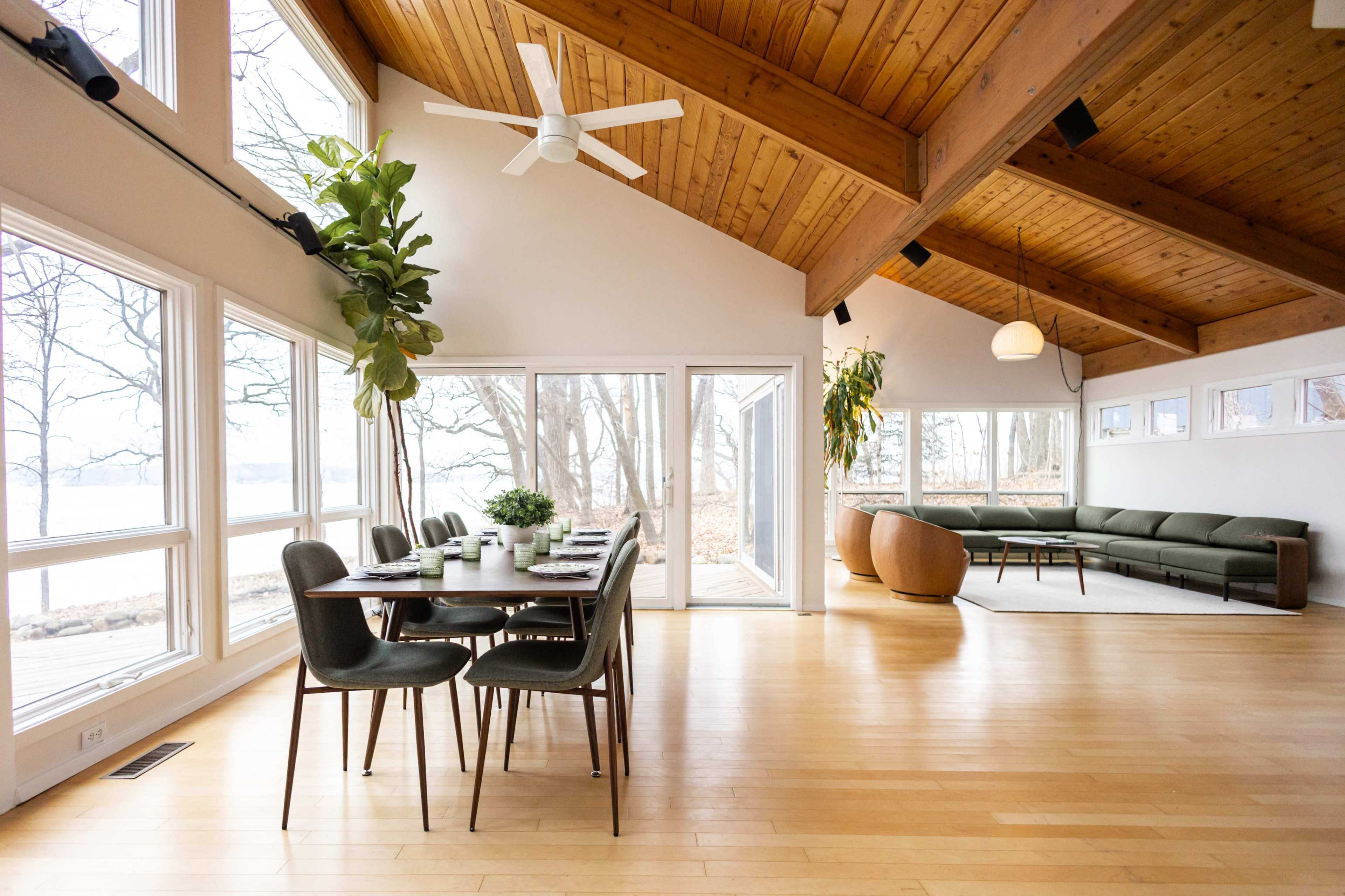 The image shows a modern interior space featuring a dining table with chairs, large windows allowing natural light to enter, and a living area with a green sofa and a round accent chair.