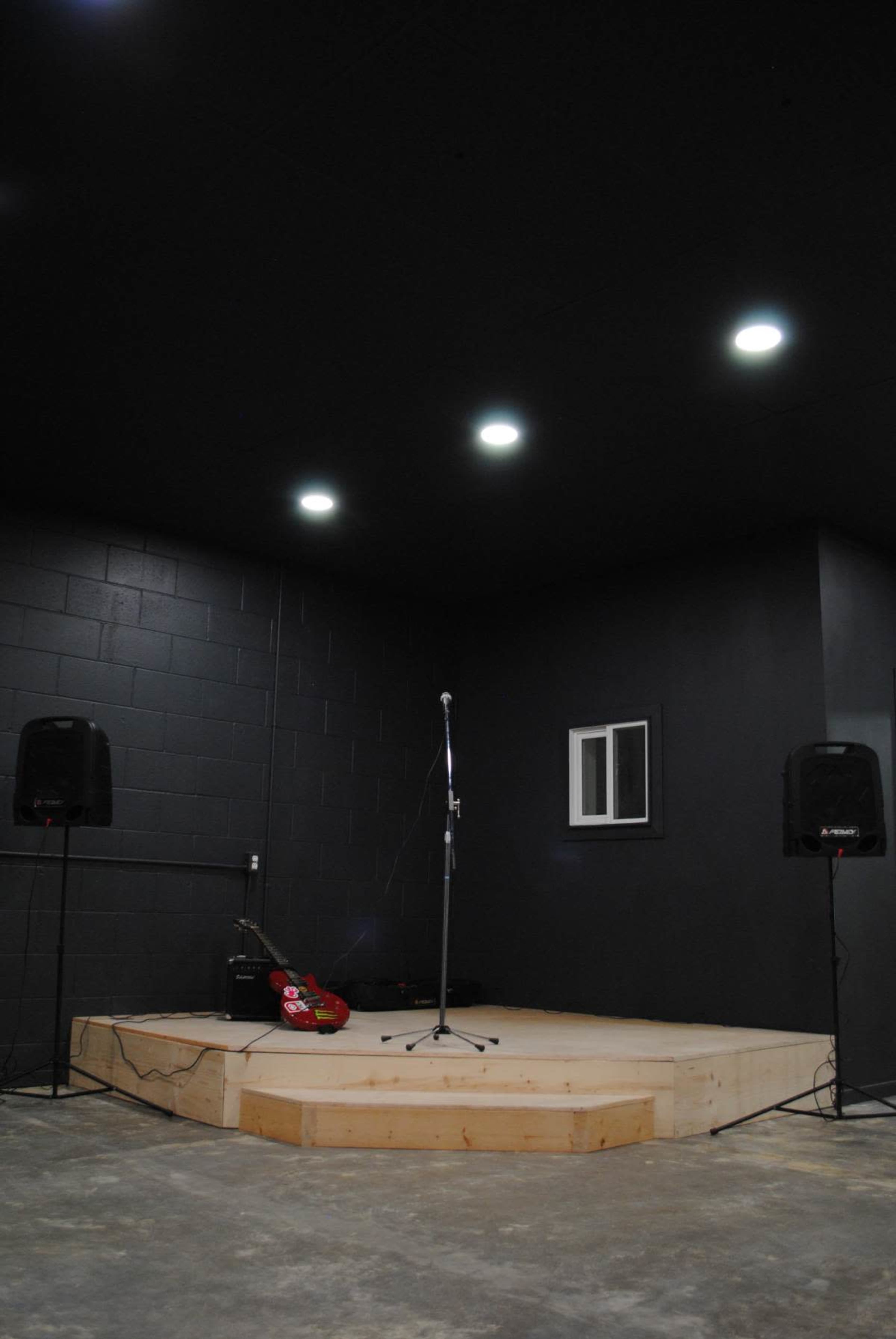 The image shows a small, elevated performance stage with a microphone set up, flanked by two speakers, in a room with black walls and a low ceiling.