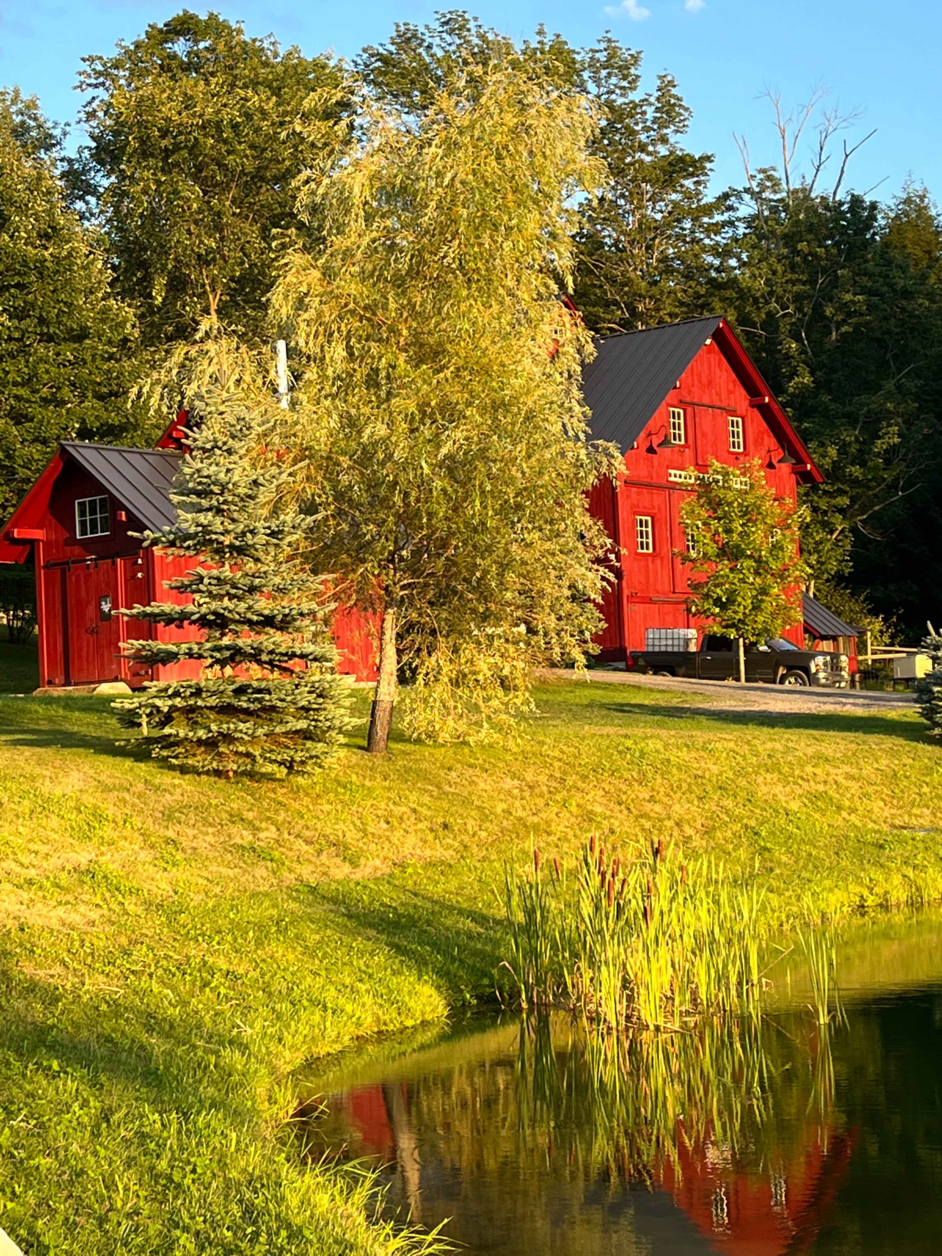 A red two-story house with a steep roof and matching outbuilding is located near a pond surrounded by trees and grass.