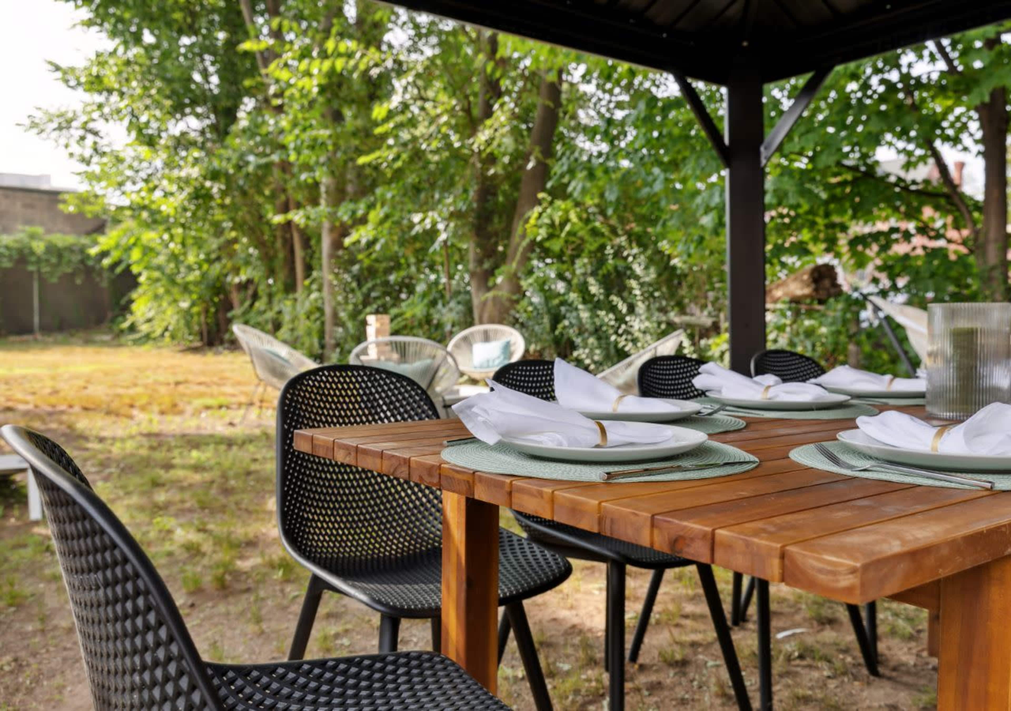 A wooden dining table is set with plates and napkins under a gazebo, surrounded by greenery in a backyard.