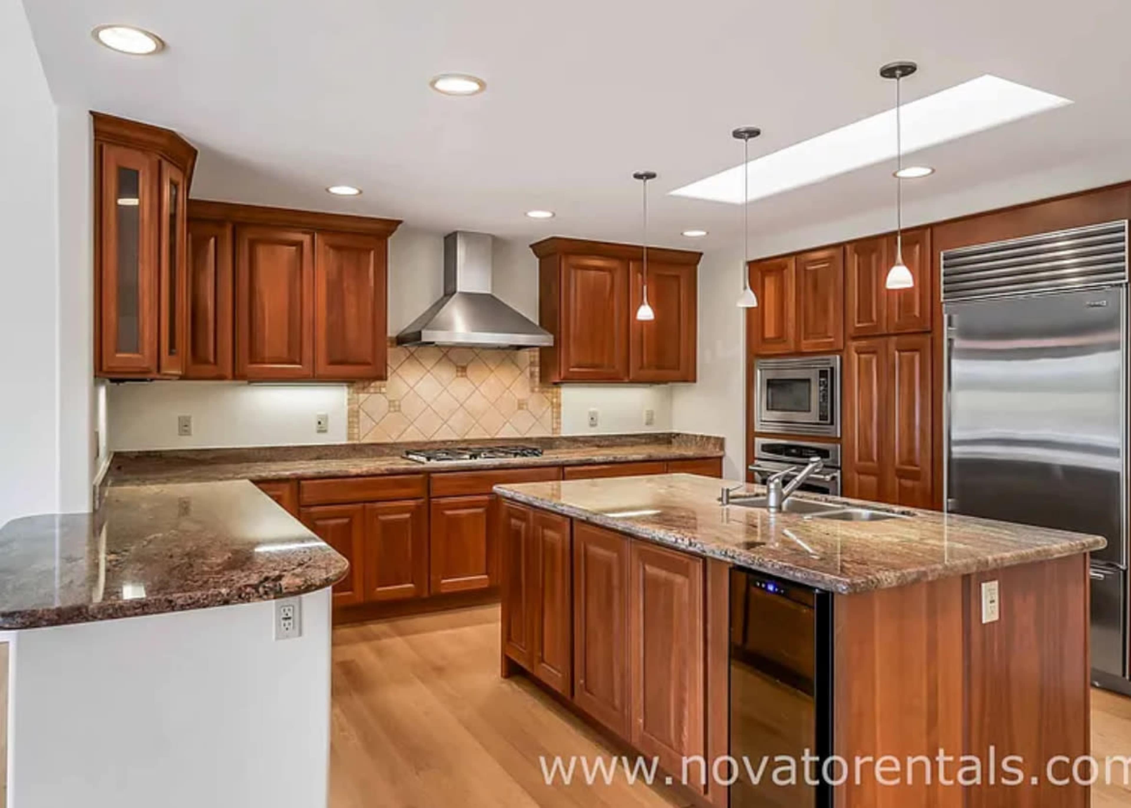 The image shows a modern kitchen featuring wooden cabinetry, stainless steel appliances, and a large island with a granite countertop.