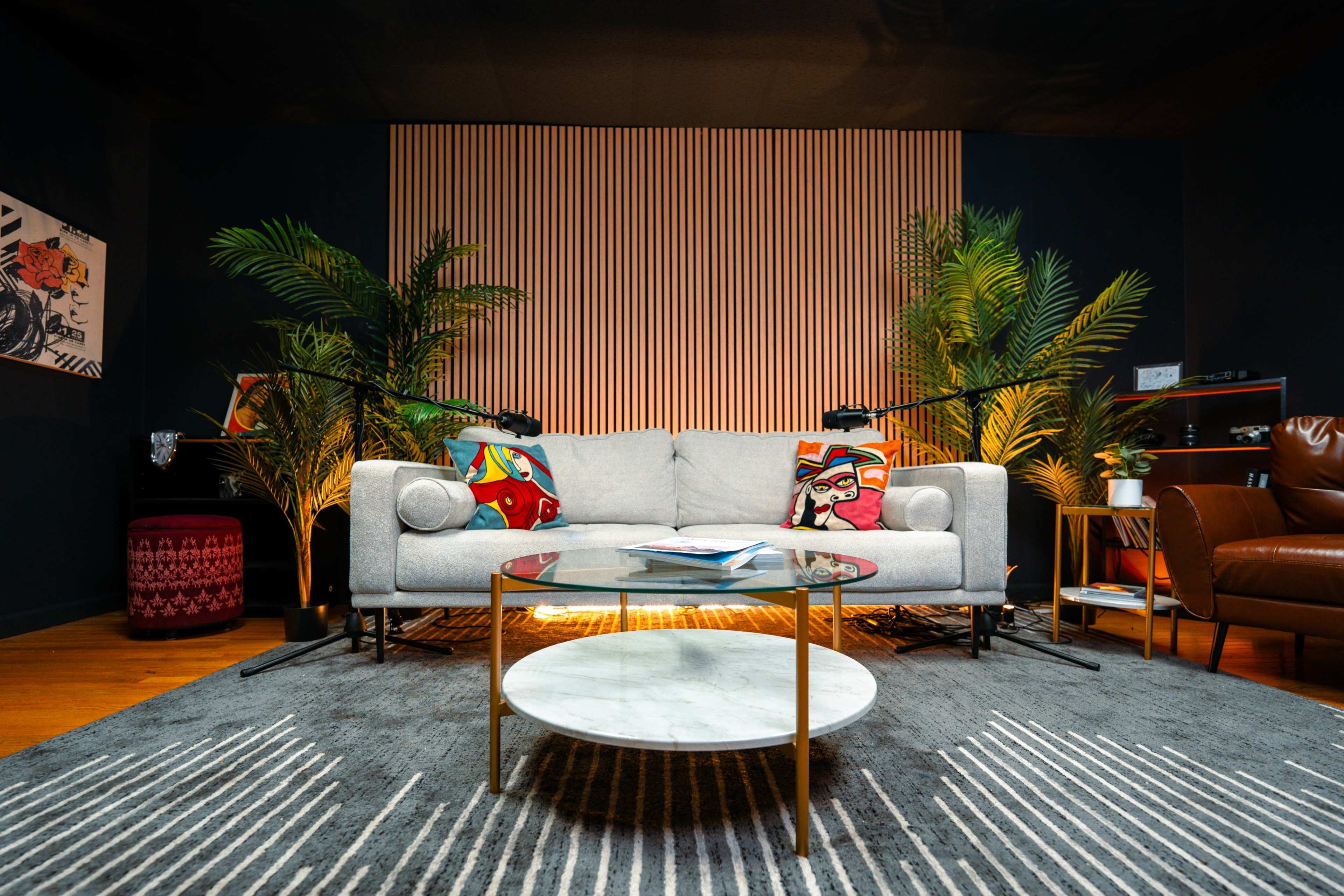 The image shows a modern living room featuring a light gray sofa with colorful cushions, a round marble coffee table, and indoor plants against a backdrop of vertical wooden slats.