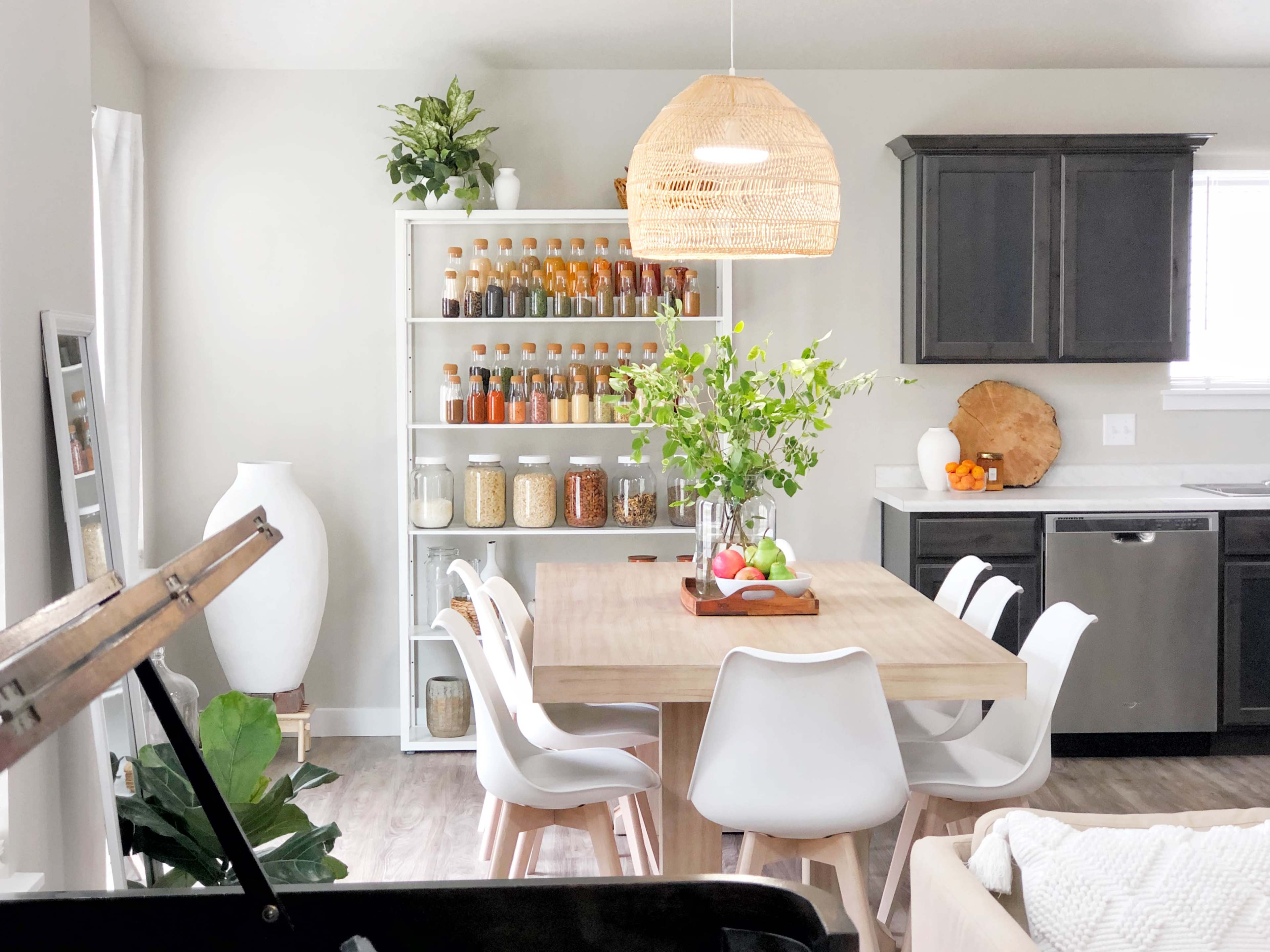 A modern kitchen features a wooden dining table surrounded by white chairs, with a shelf of jars and a pendant light overhead.