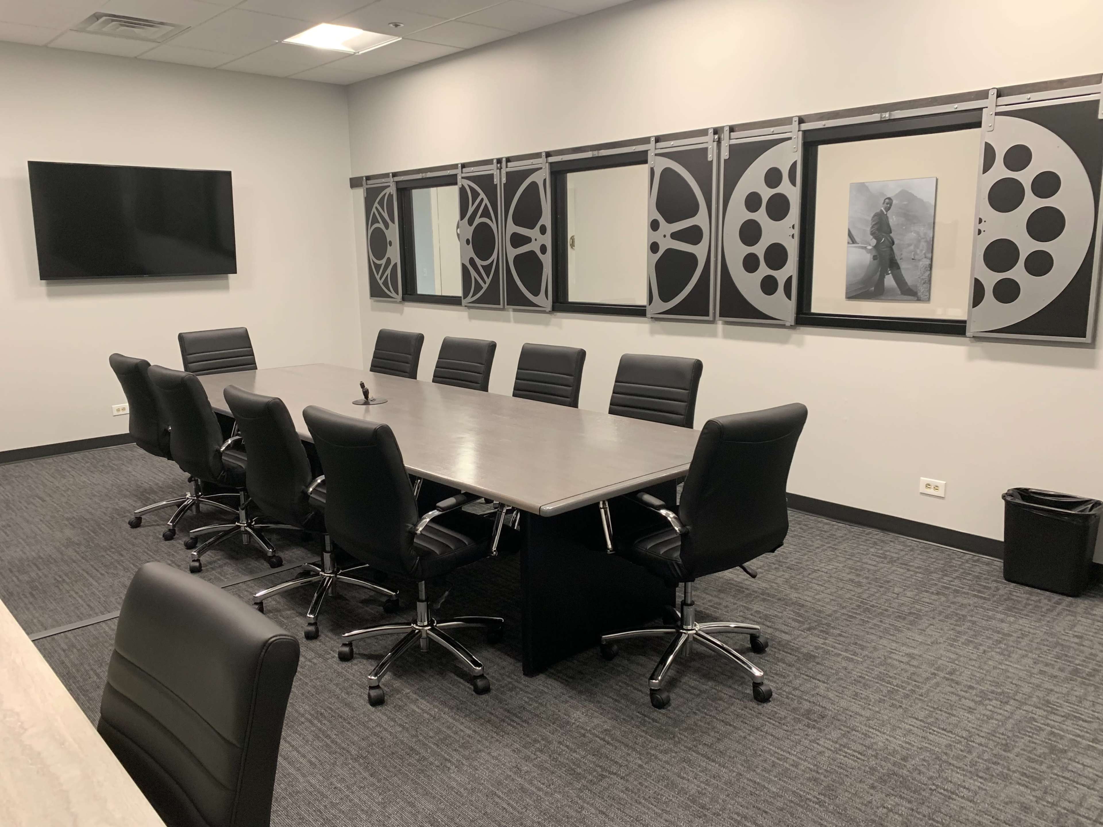 A conference room with a long rectangular table surrounded by ten black leather chairs, a large screen on the wall, and film reel-themed decor.