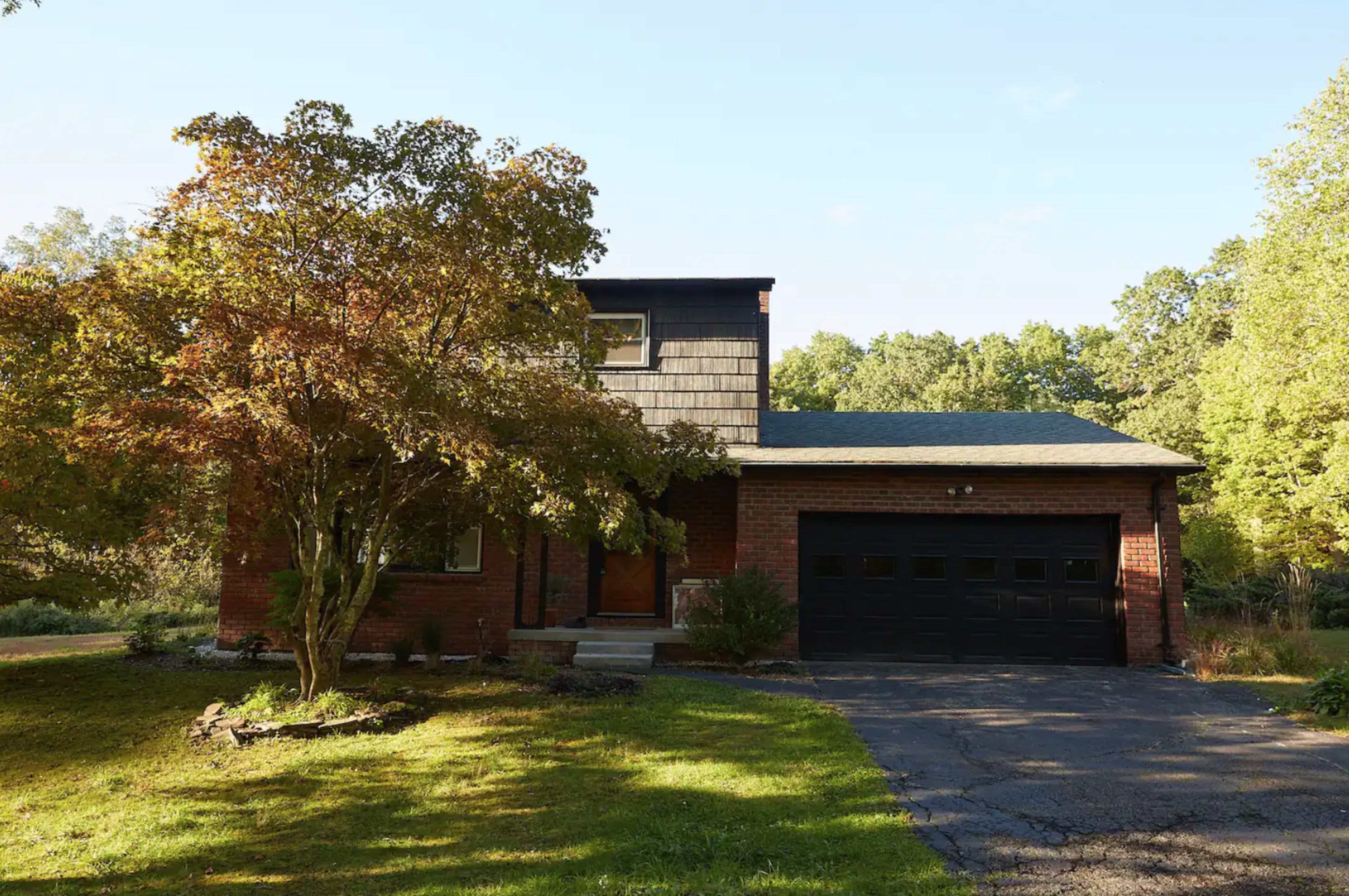 A two-story brick house with a dark shingle roof and a garage is surrounded by greenery and a well-maintained lawn.