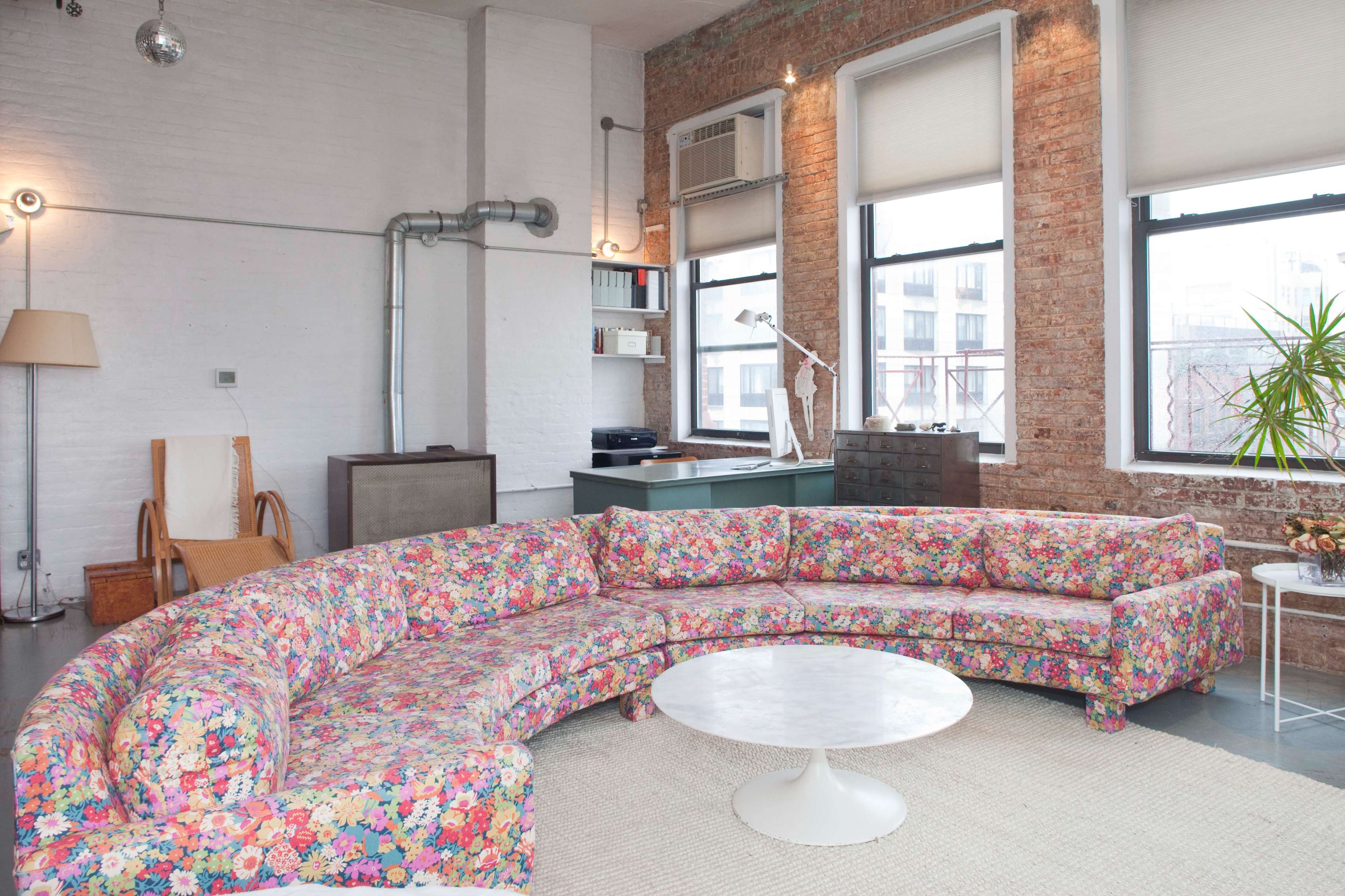 A living room with a curved floral-patterned couch, a round white coffee table, and large windows that let in natural light.