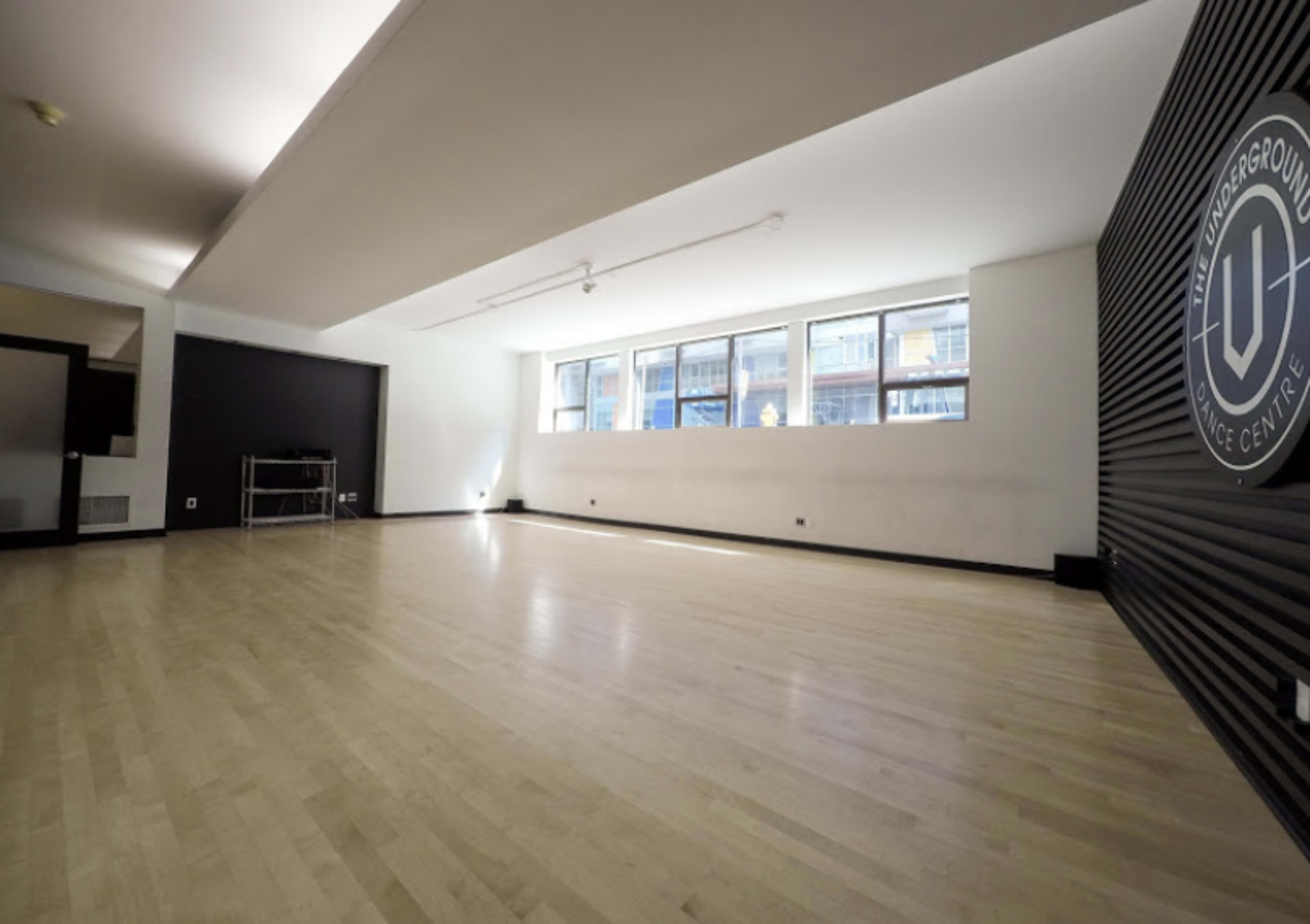 A spacious, empty room with wooden flooring and large windows, featuring a black wall with a circular logo.